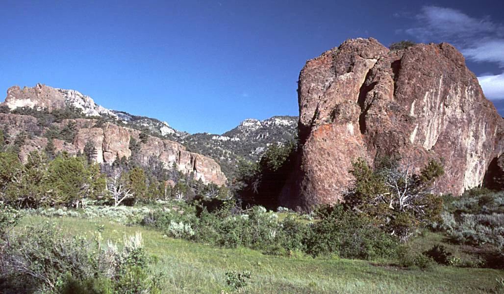 A large, weathered brown rock monolith stands prominently on the right side of the frame, with expansive cliff faces and distant mountains forming a rugged backdrop over a grassy, sagebrush-covered valley.