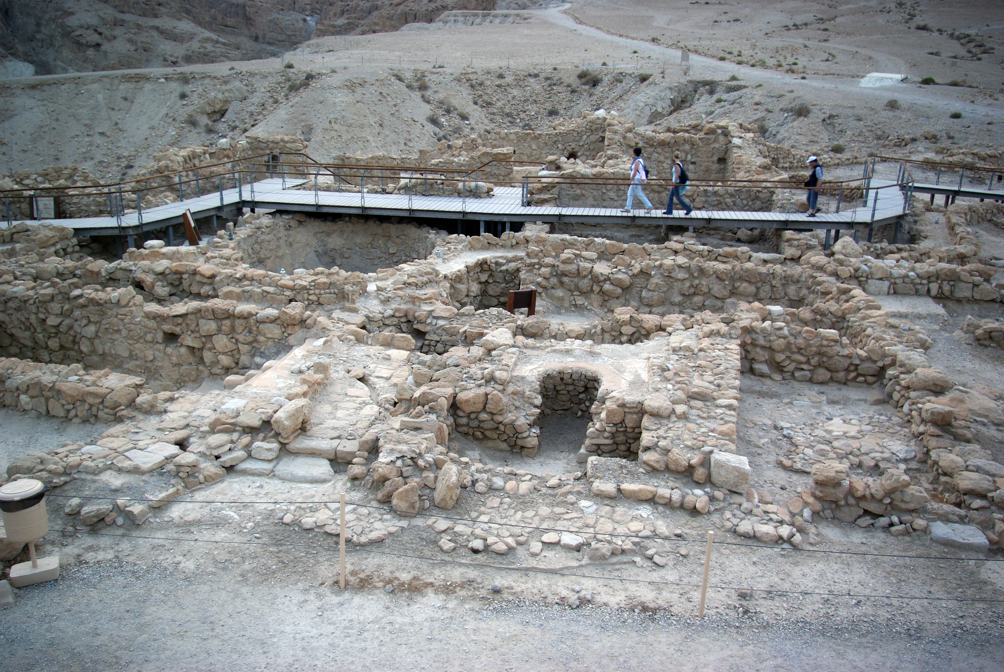 Black and white photograph showing ancient stone ruins with a wooden boardwalk, several people walking, and desert terrain in the background