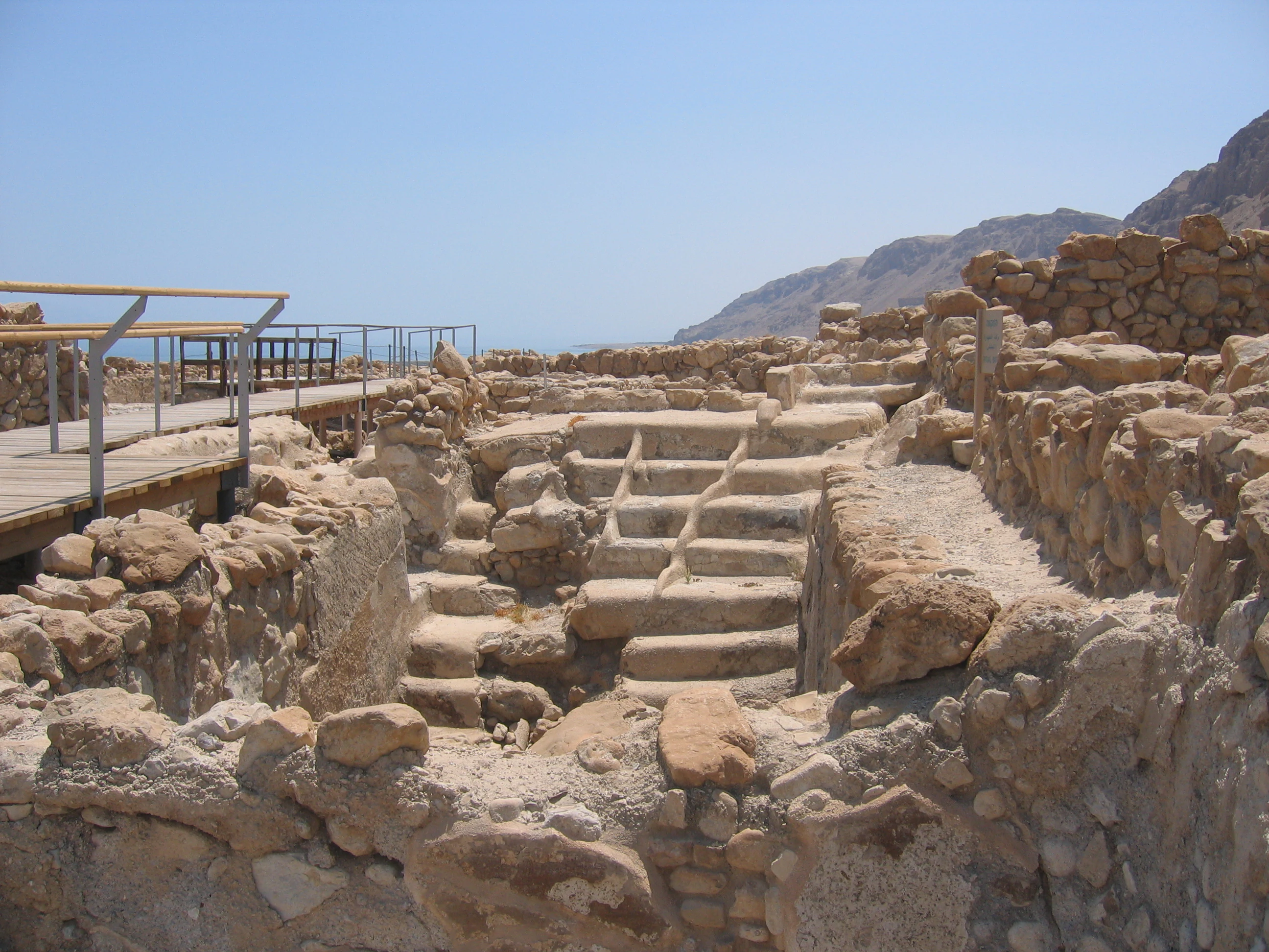 Stone ritual bath structure with steps surrounded by ancient stone walls, wooden walkway with railings, mountains and Dead Sea visible in background