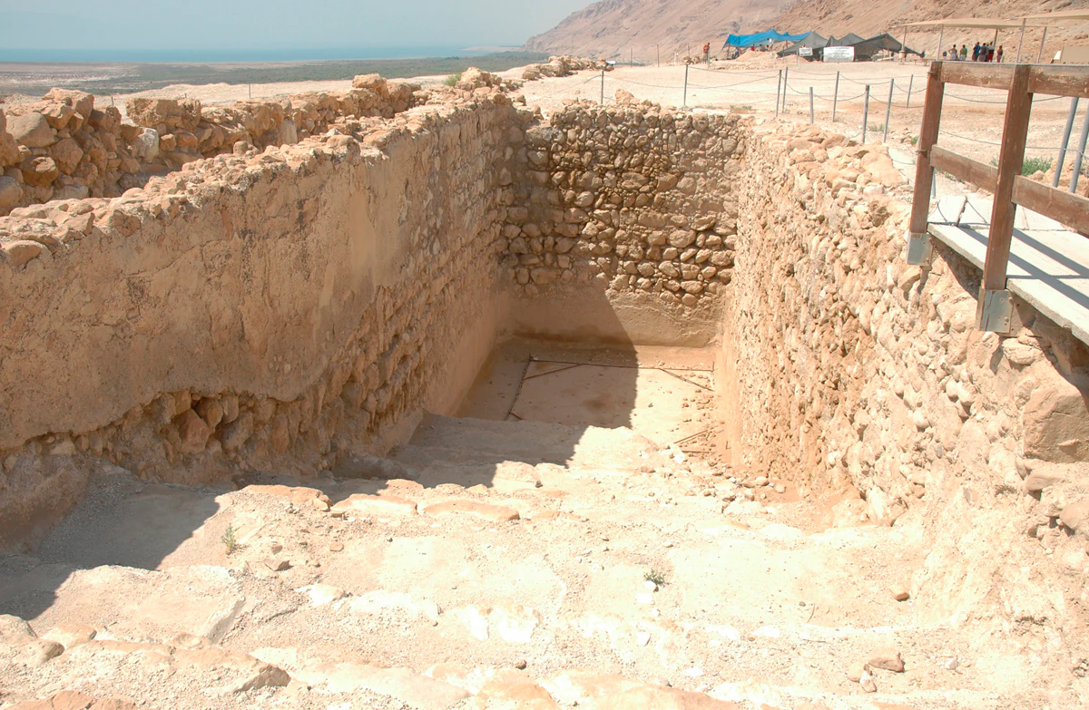 Stone steps descending into a rectangular ancient pool with stone walls, wooden railing on the right, and desert landscape in the background