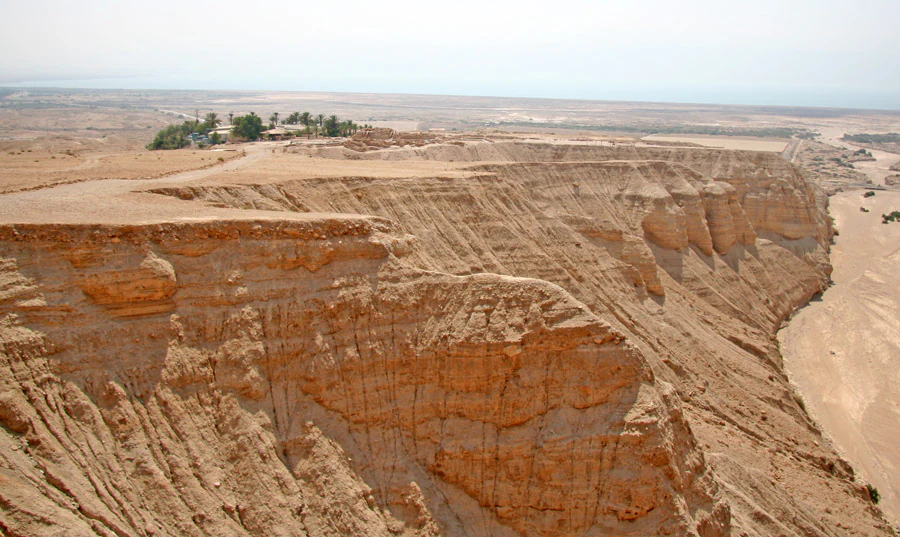 Desert landscape with rocky cliffs, sandy terrain, and the Dead Sea visible in the distance