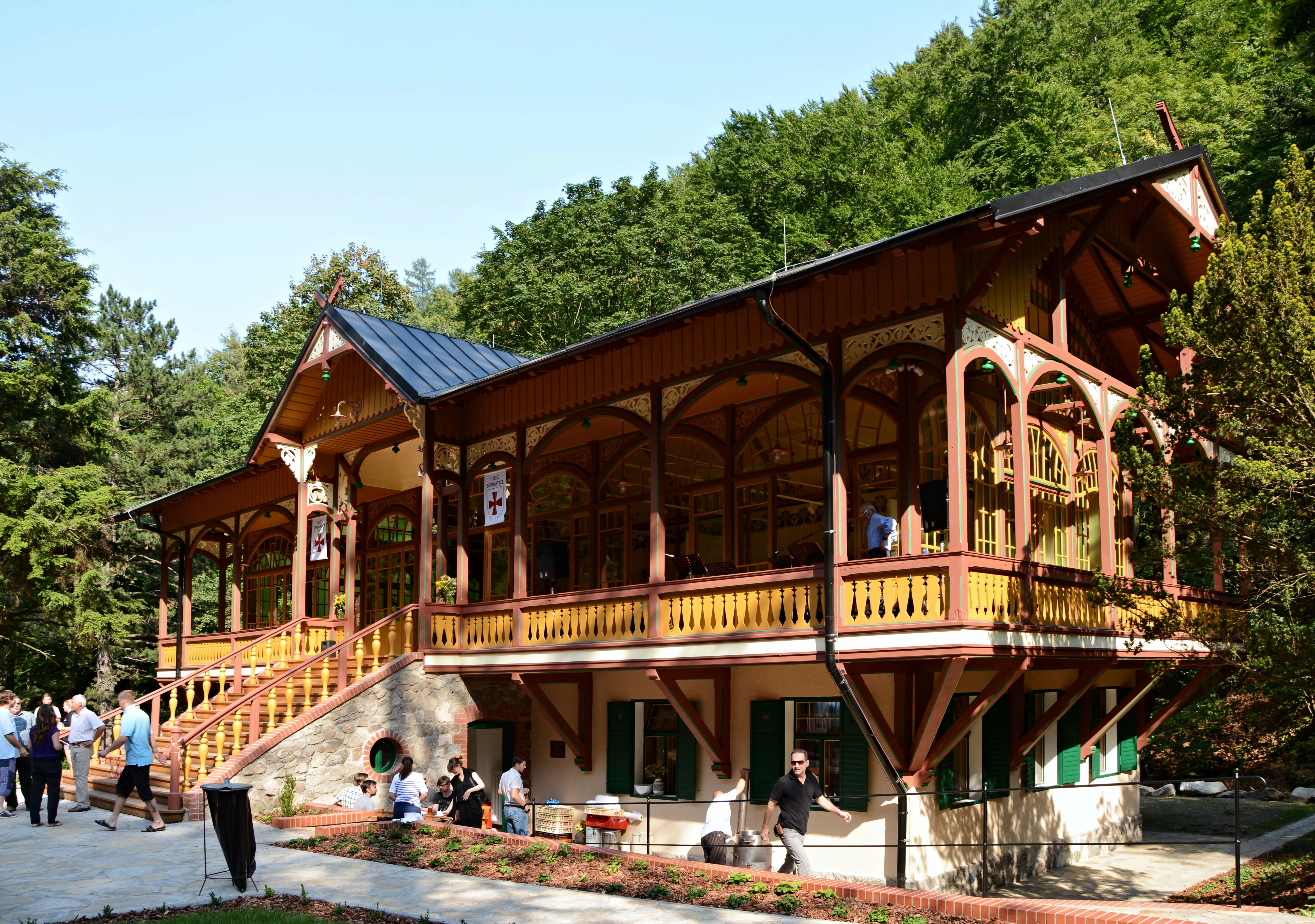 A restored wooden ballroom building with ornate timber framing and a stone foundation, set against a forest backdrop with several visitors walking on the plaza in front.