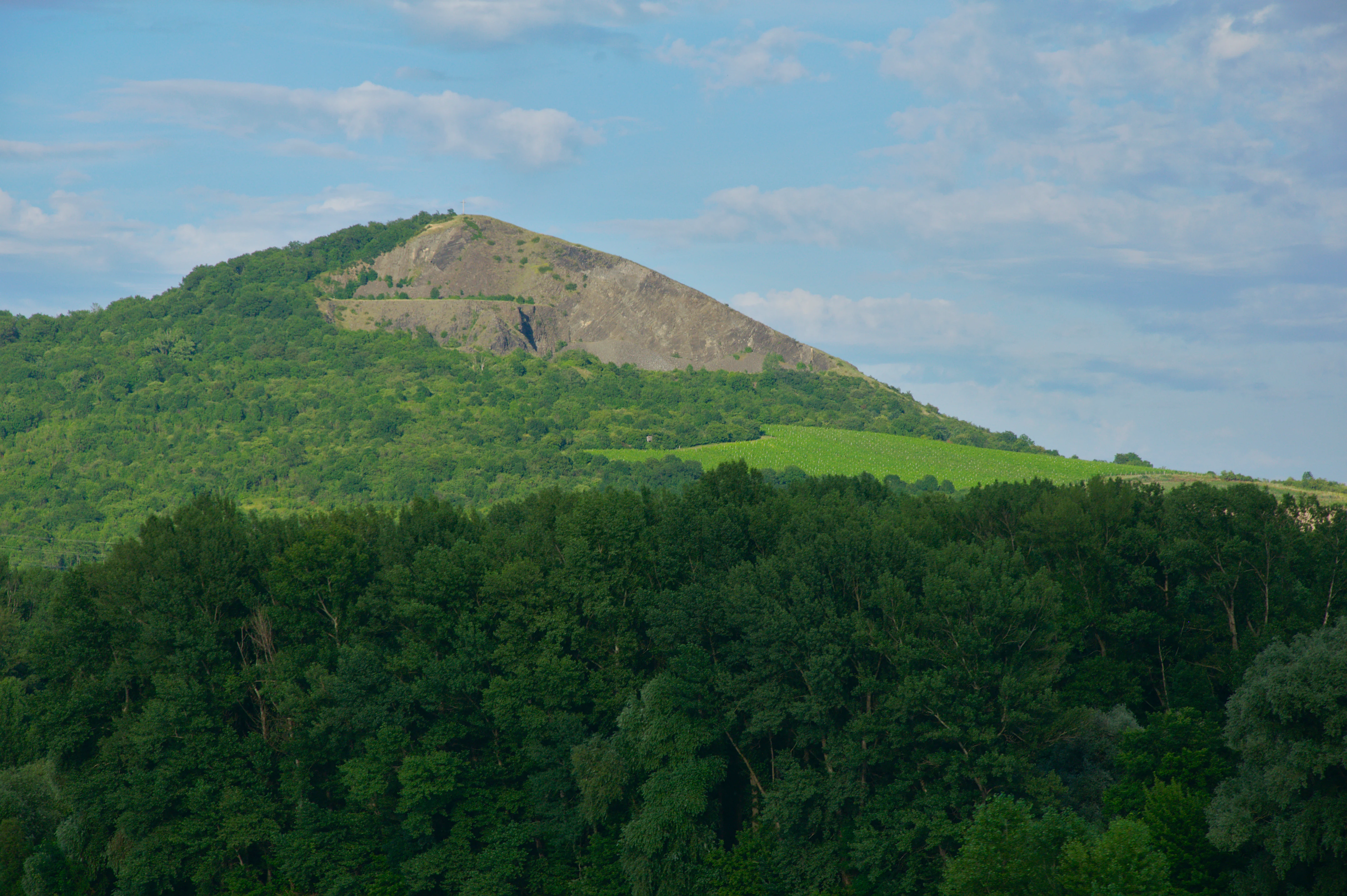 A green forested hill with a visible rocky, basaltic peak rising against a partly cloudy sky.