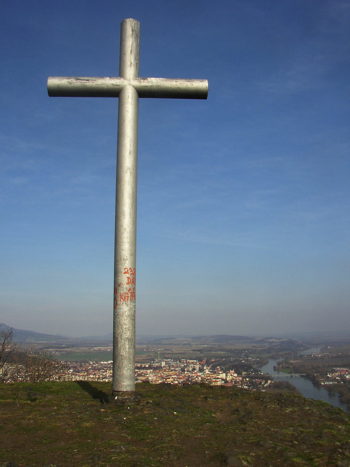 A tall, weathered metal cross stands prominently on a grassy hill summit against a blue sky, with a town and river visible in the distance.