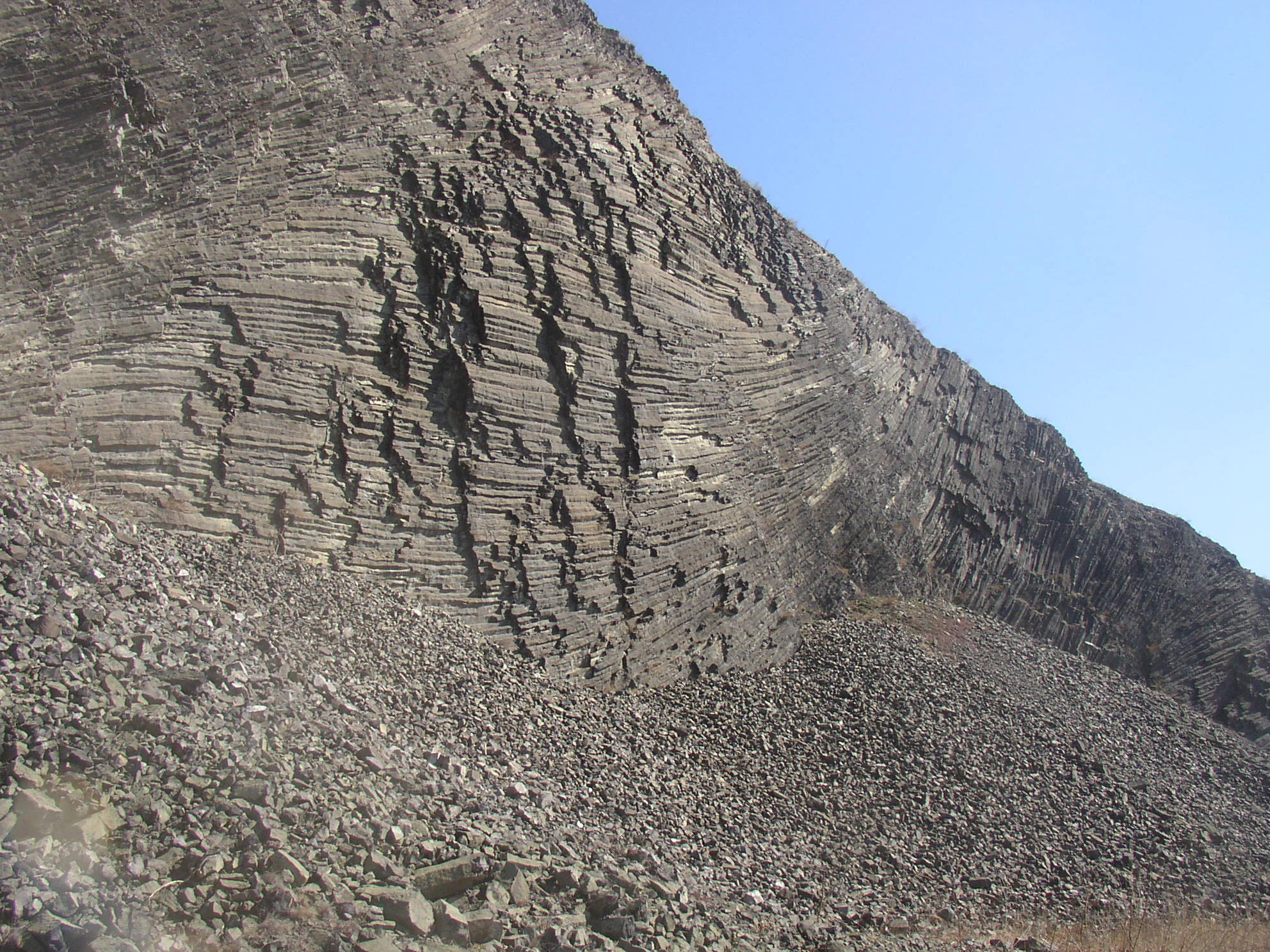 A close-up view of a steep cliff face featuring weathered basalt columns in an abandoned quarry, with a large scree slope of loose rock below.