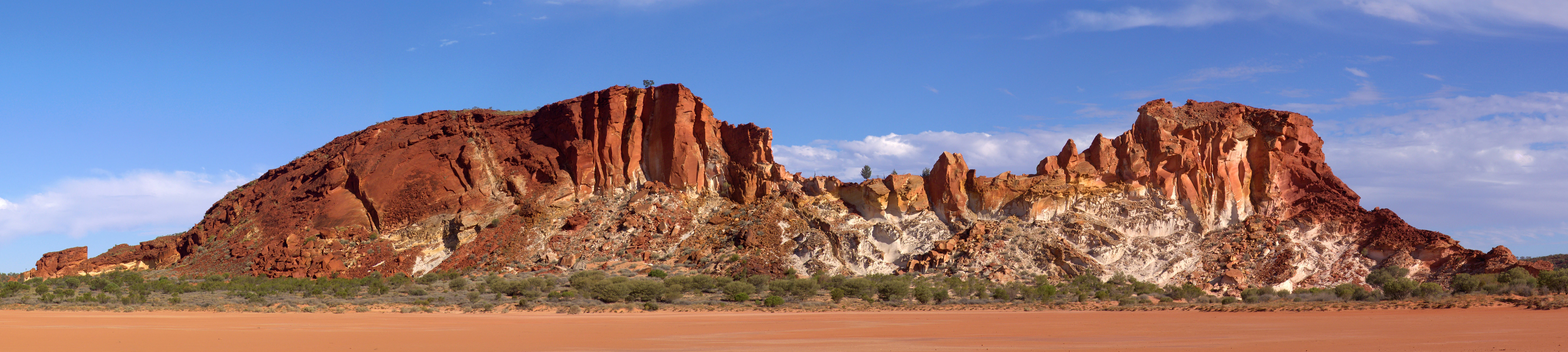 A panoramic wide shot of red sandstone bluffs with vertical striations, rising above a flat, sandy desert floor under a clear blue sky.