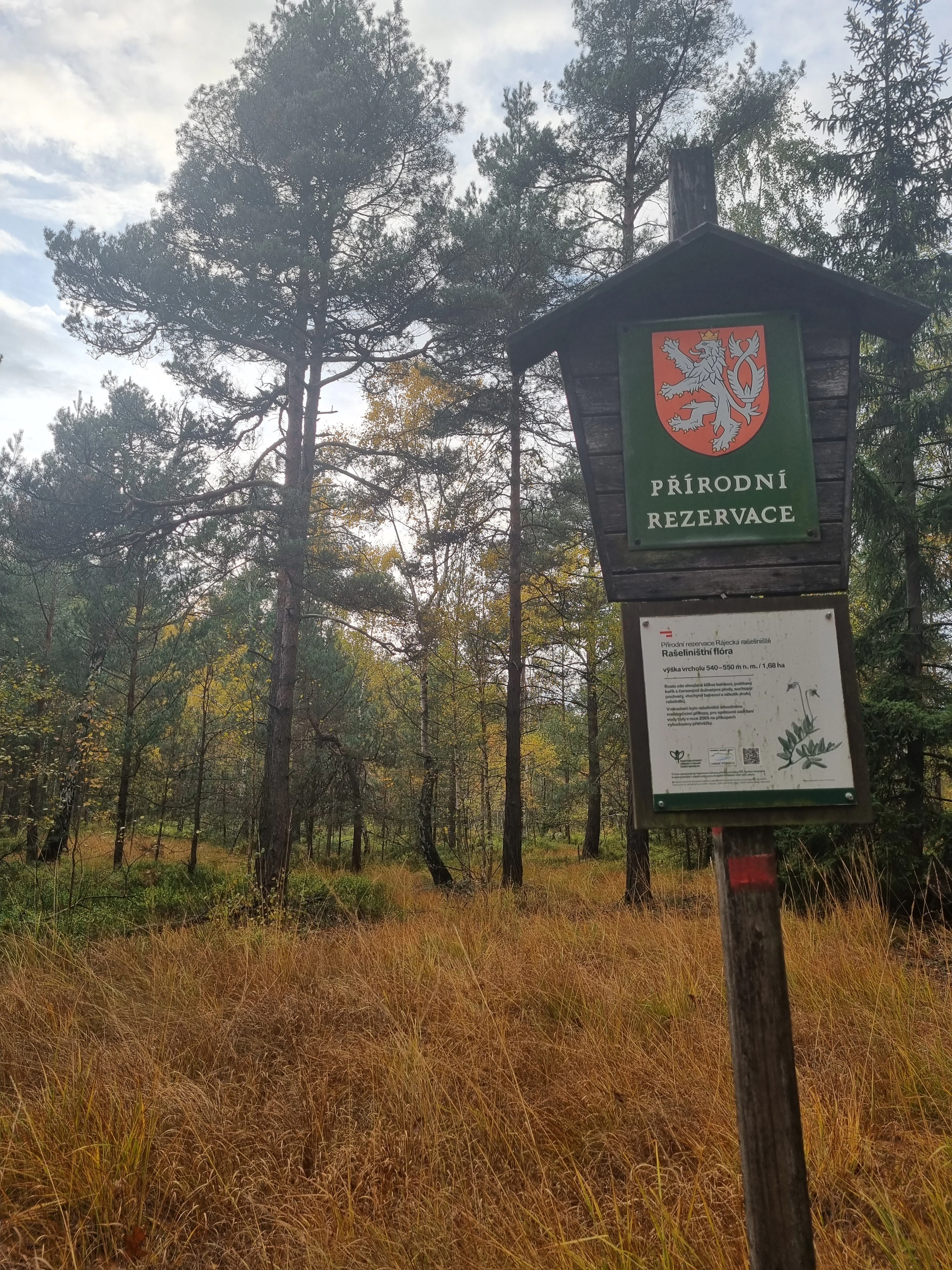 A wooden informational signpost with the Czech lion crest labeled PŘÍRODNÍ REZERVACE stands in a grassy, forested area.