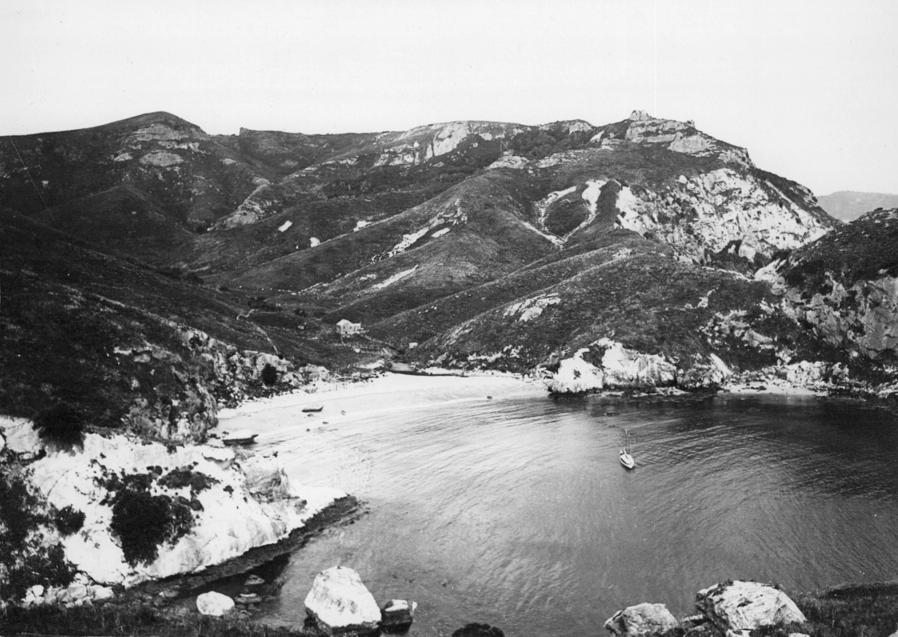 A black and white archival photograph showing a protected coastal bay on Rakitu Island, with steep rocky hills rising above a sandy beach and calm water where a small boat is visible.