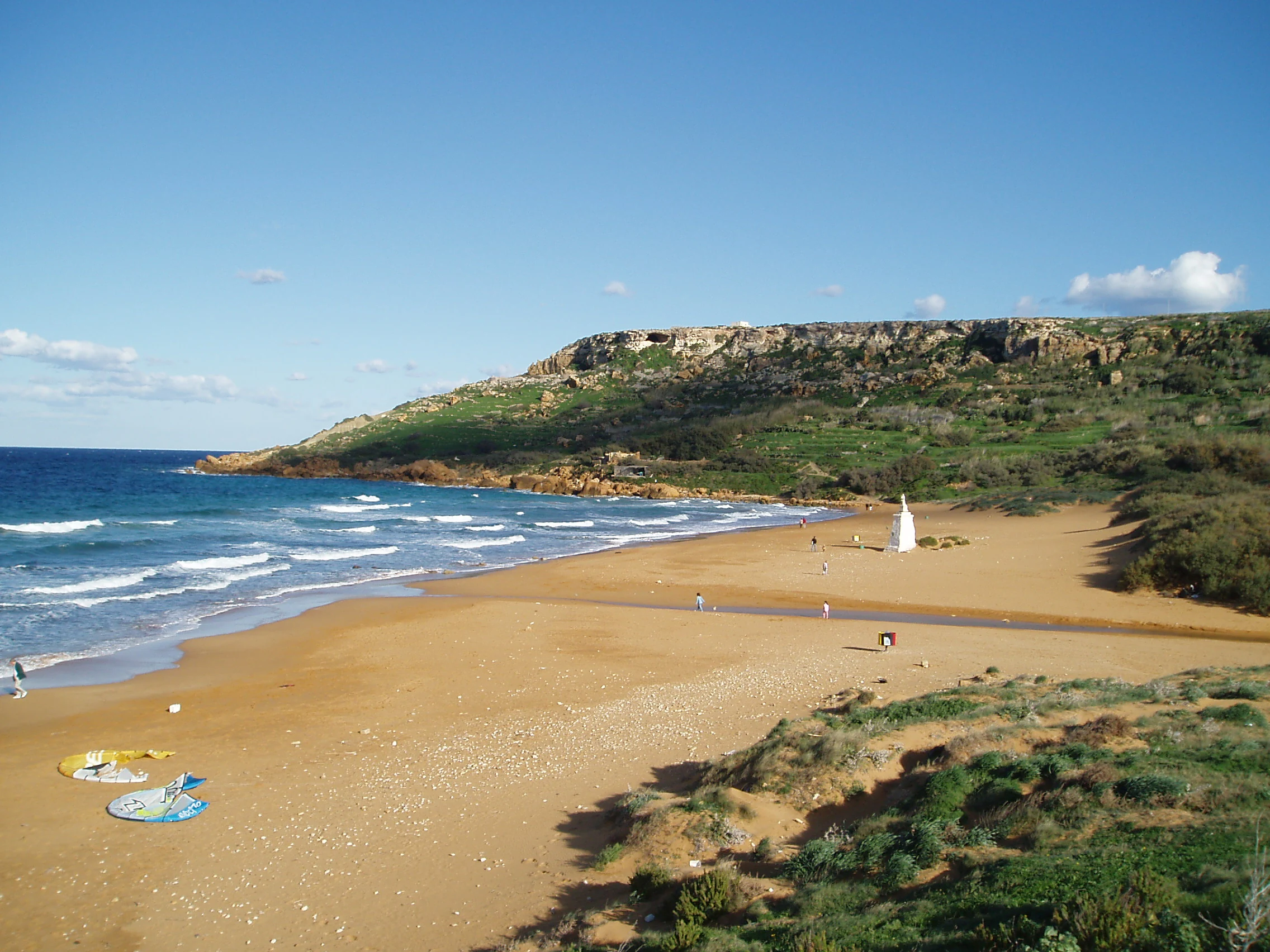 A wide coastal landscape showing a sandy beach with reddish-hued sand meeting the blue sea, framed by a prominent rocky cliff in the background and low dunes in the foreground.