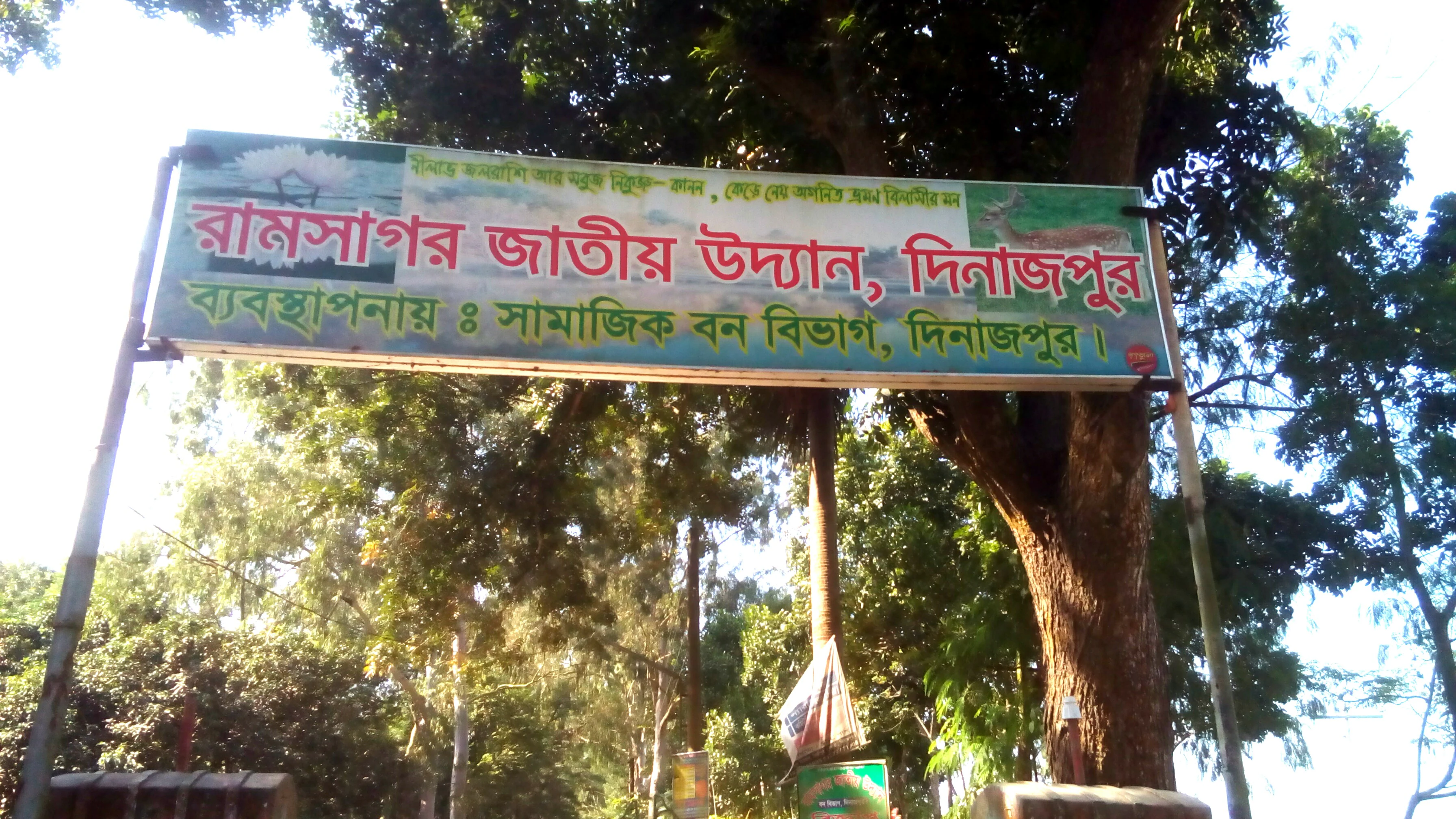 Cartel de entrada verde y blanco con texto en bengalí, estructura de puerta metálica y árboles a su alrededor en la entrada del Parque Nacional Ramsagar