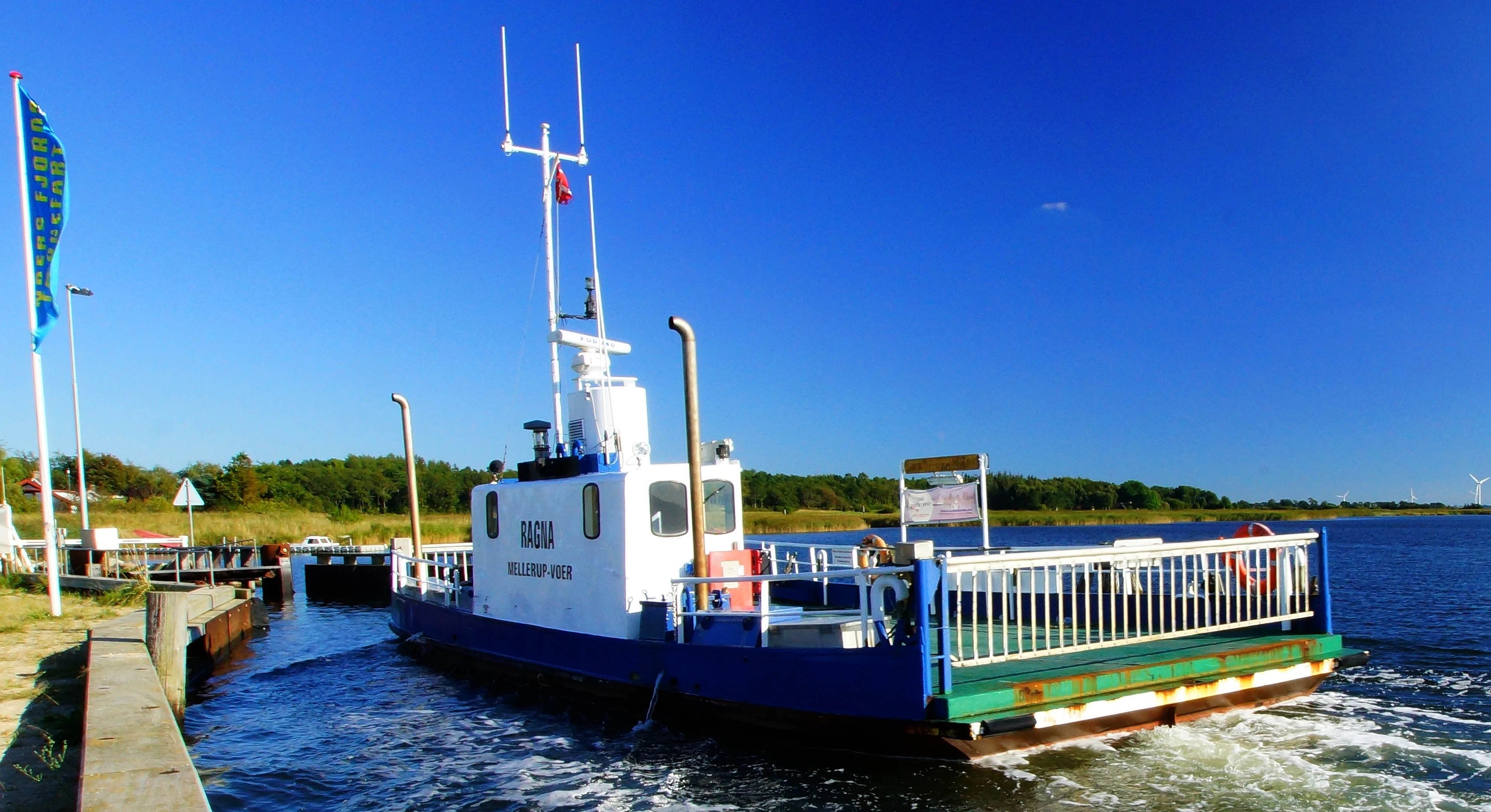 A small white and blue ferry named Ragna docked at a pier on Randers Fjord under a clear blue sky.