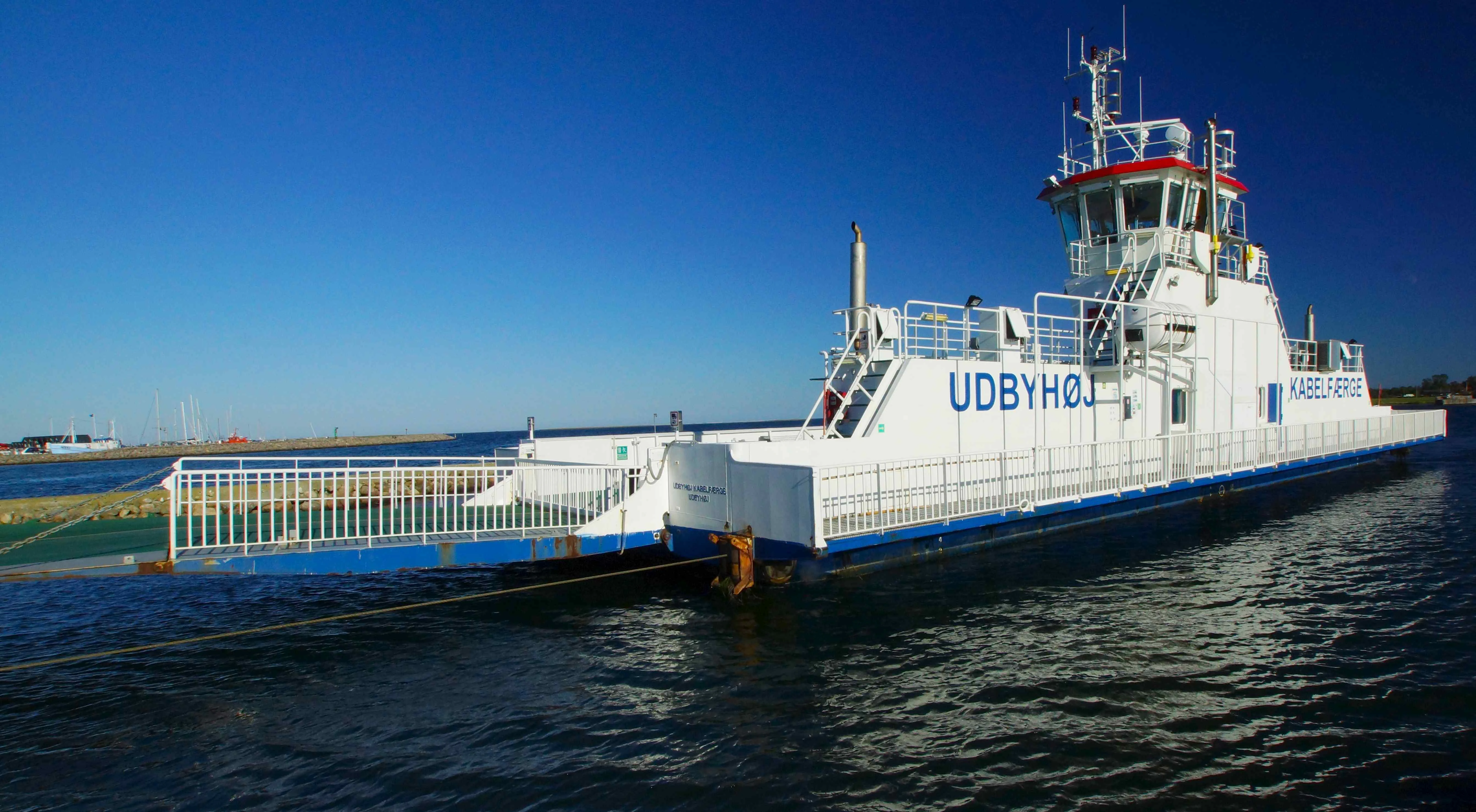 A white and blue cable ferry named Udbyhoj sits on calm blue water under a clear sky.