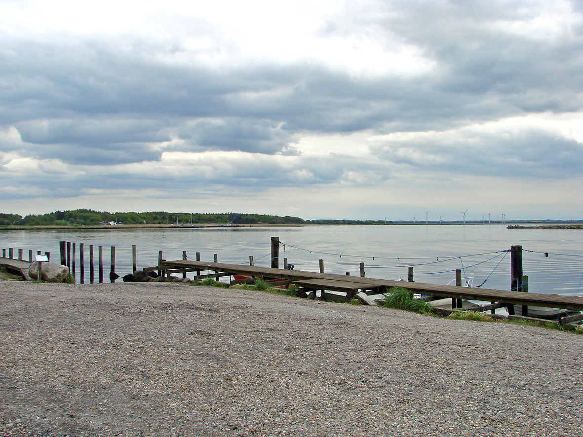 A gravel foreground leads to a wooden pier extending into the calm waters of Randers Fjord under a cloudy sky.