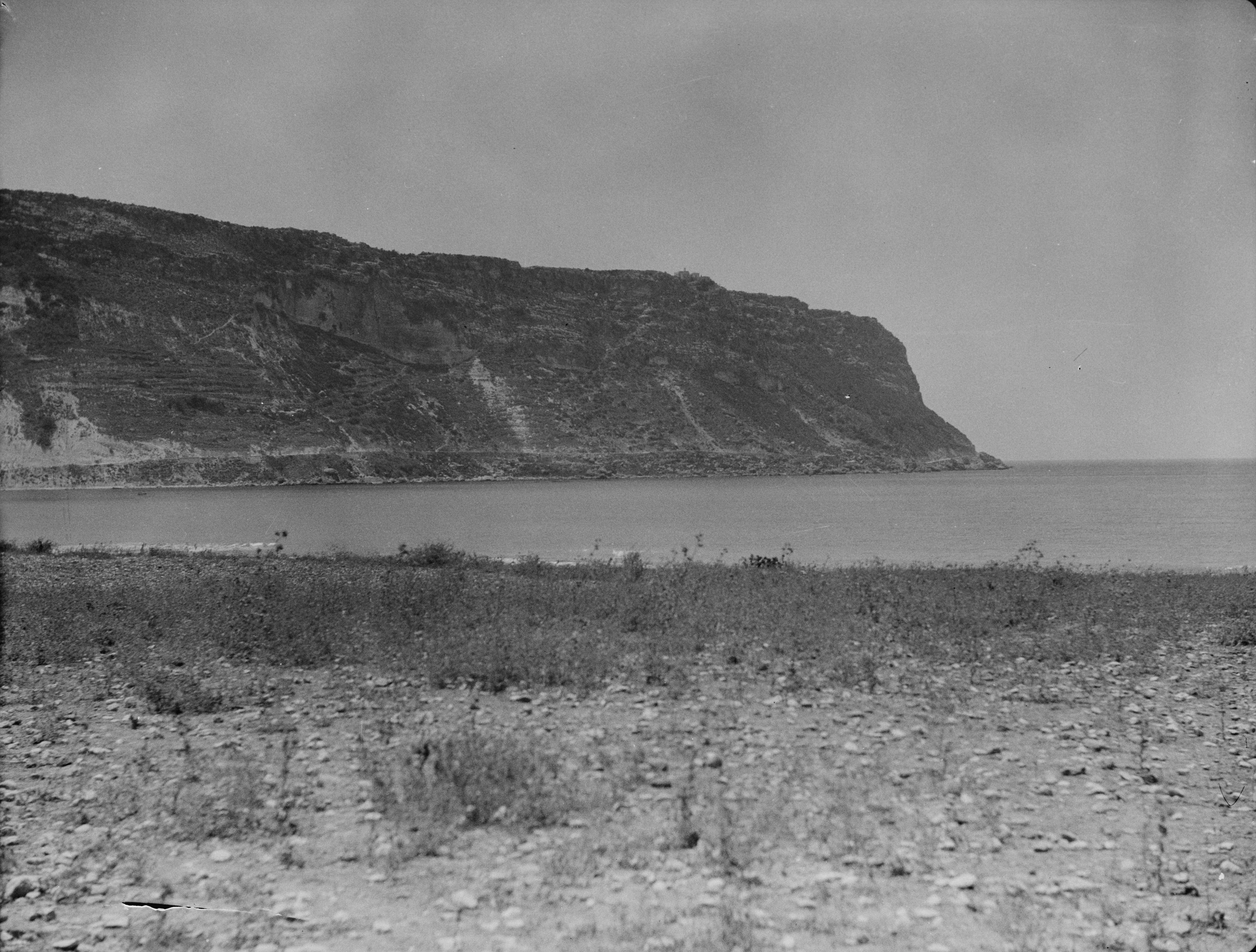 A black and white archival photograph showing a prominent coastal cliff or headland extending into the sea, with a rocky and sparsely vegetated shoreline in the foreground.