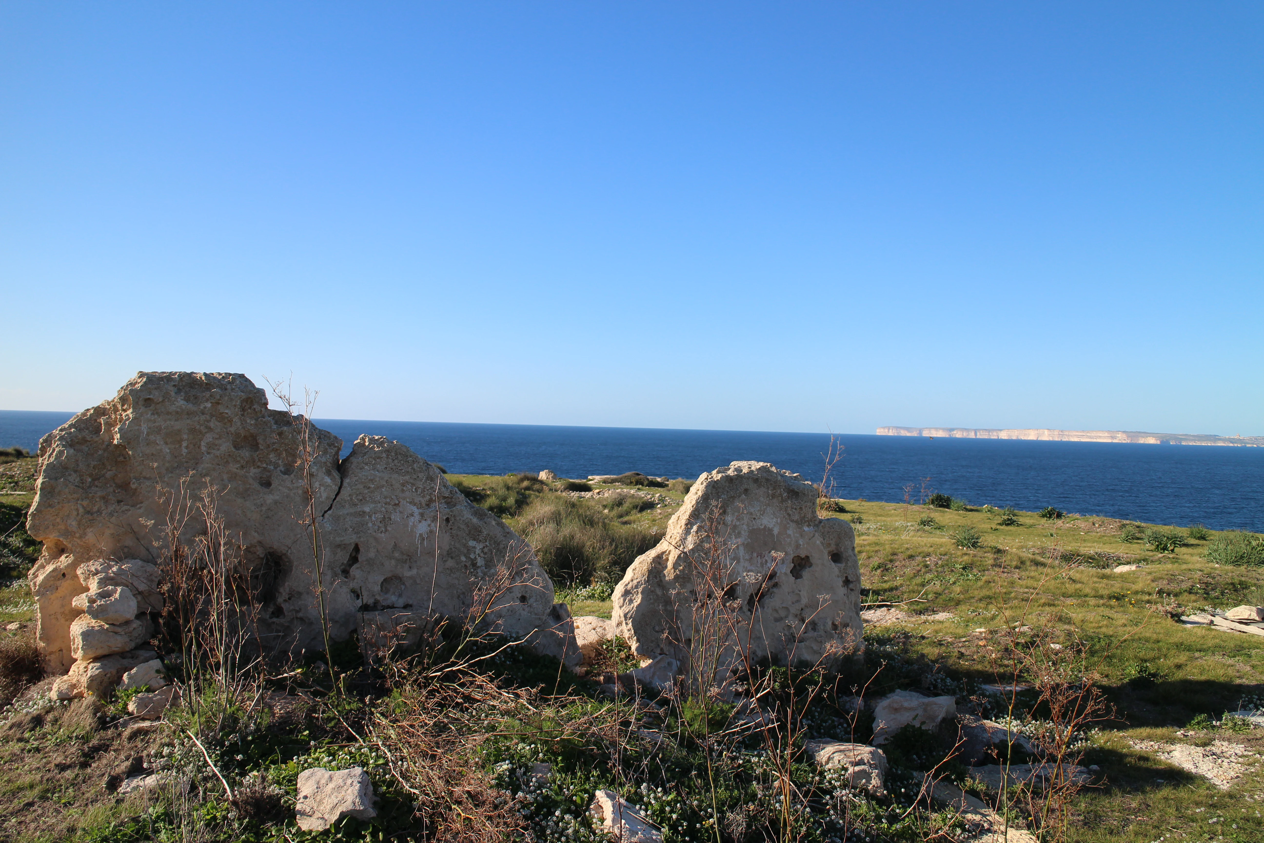 Ancient stone ruins of a megalithic structure situated on a grassy cliff overlooking the blue Mediterranean Sea on a clear day.