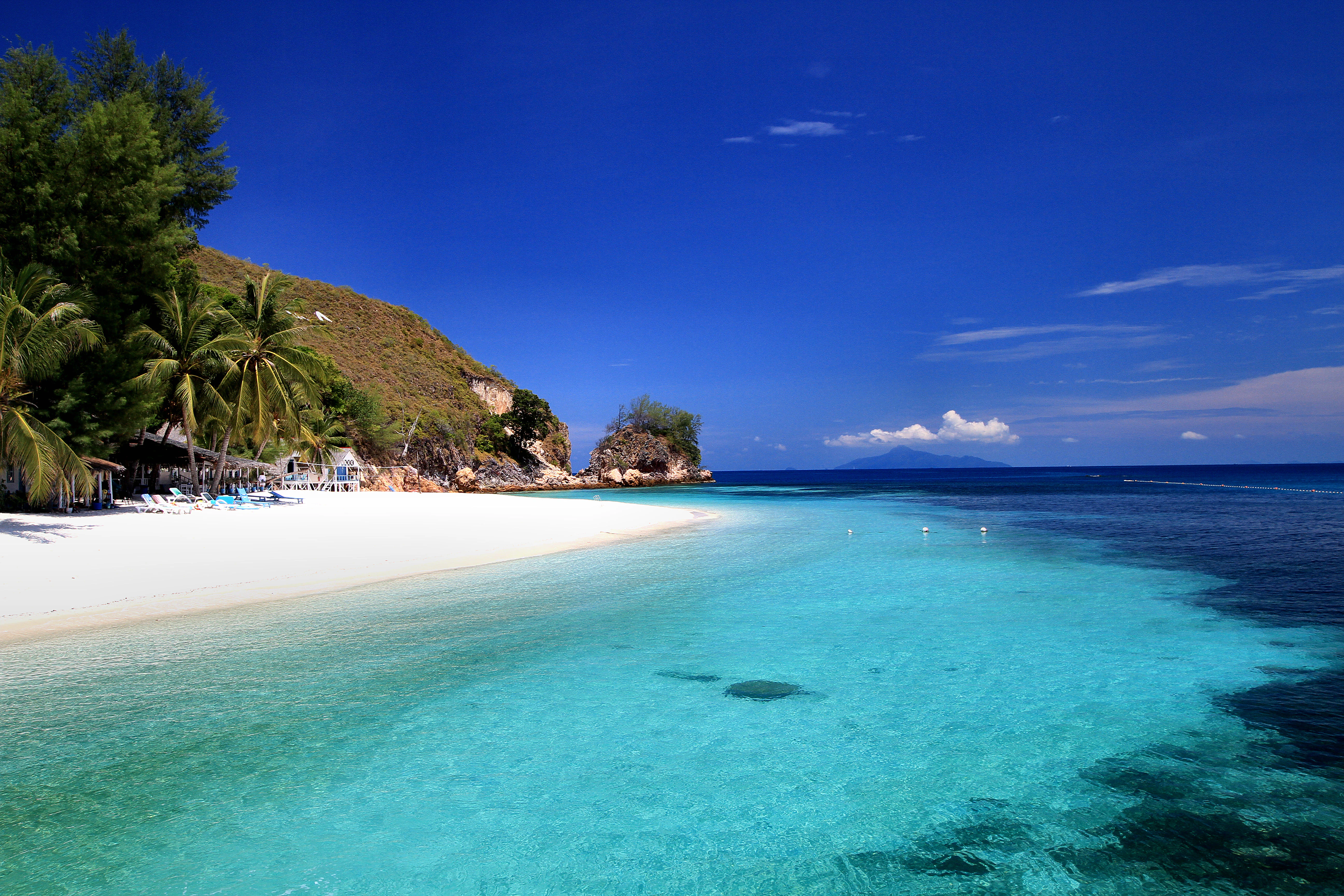 A wide angle view of a pristine white sandy beach bordering clear turquoise shallow water, with a rocky hillside and palm trees on the left under a deep blue sky.