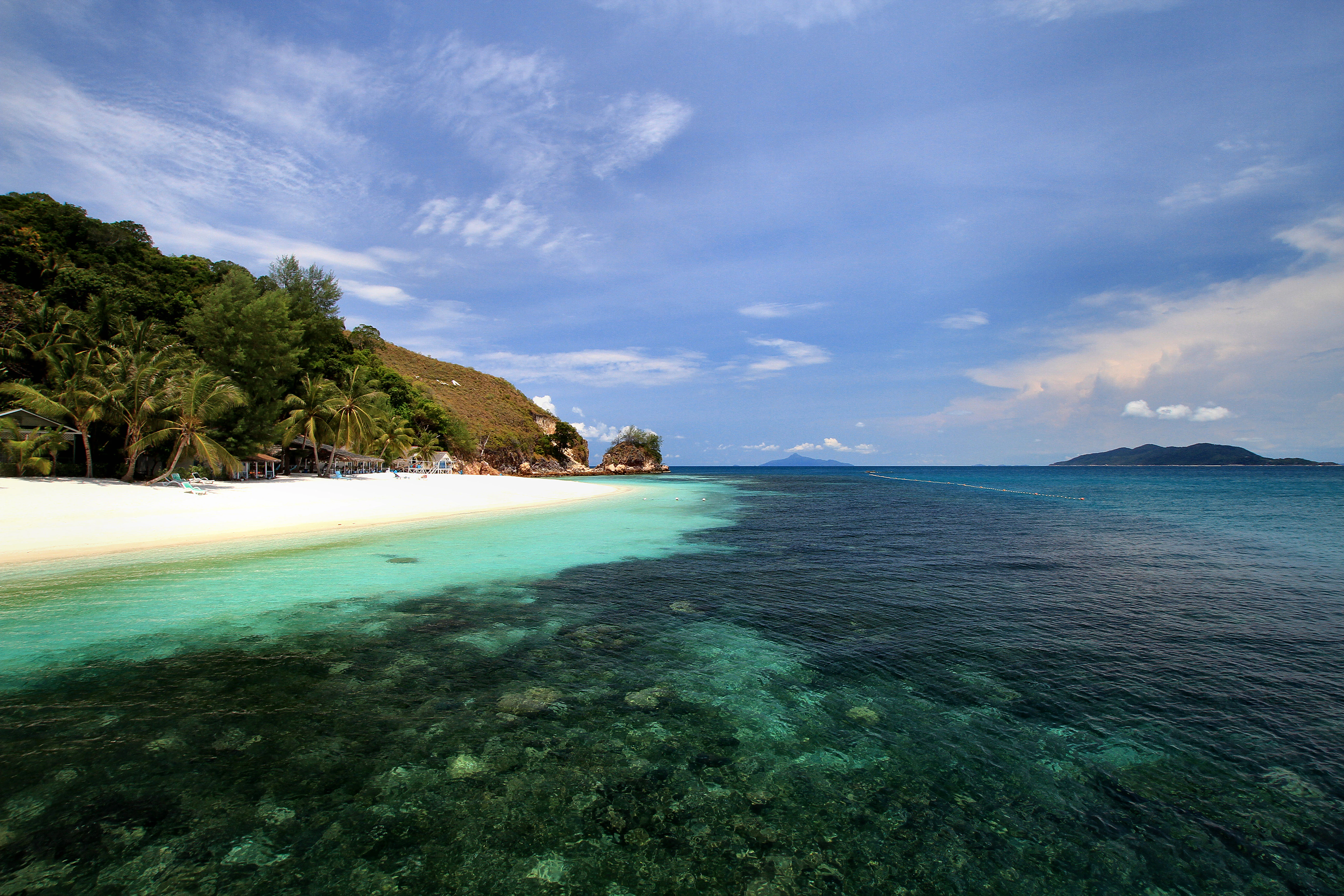 A wide angle view of a white sand beach meeting clear turquoise water on Rawa Island, with lush green hills in the background under a blue sky.