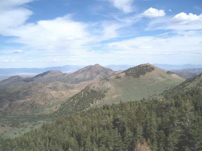 A wide, high-angle shot showing rolling reddish-orange volcanic mountains and pine forests under a bright blue sky with scattered clouds.