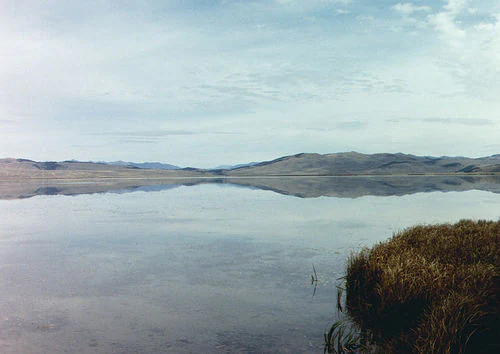A wide, eye level landscape shot of a calm, reflective lake under a pale blue sky, with grassy marshes in the foreground and rounded, distant mountains across the horizon.