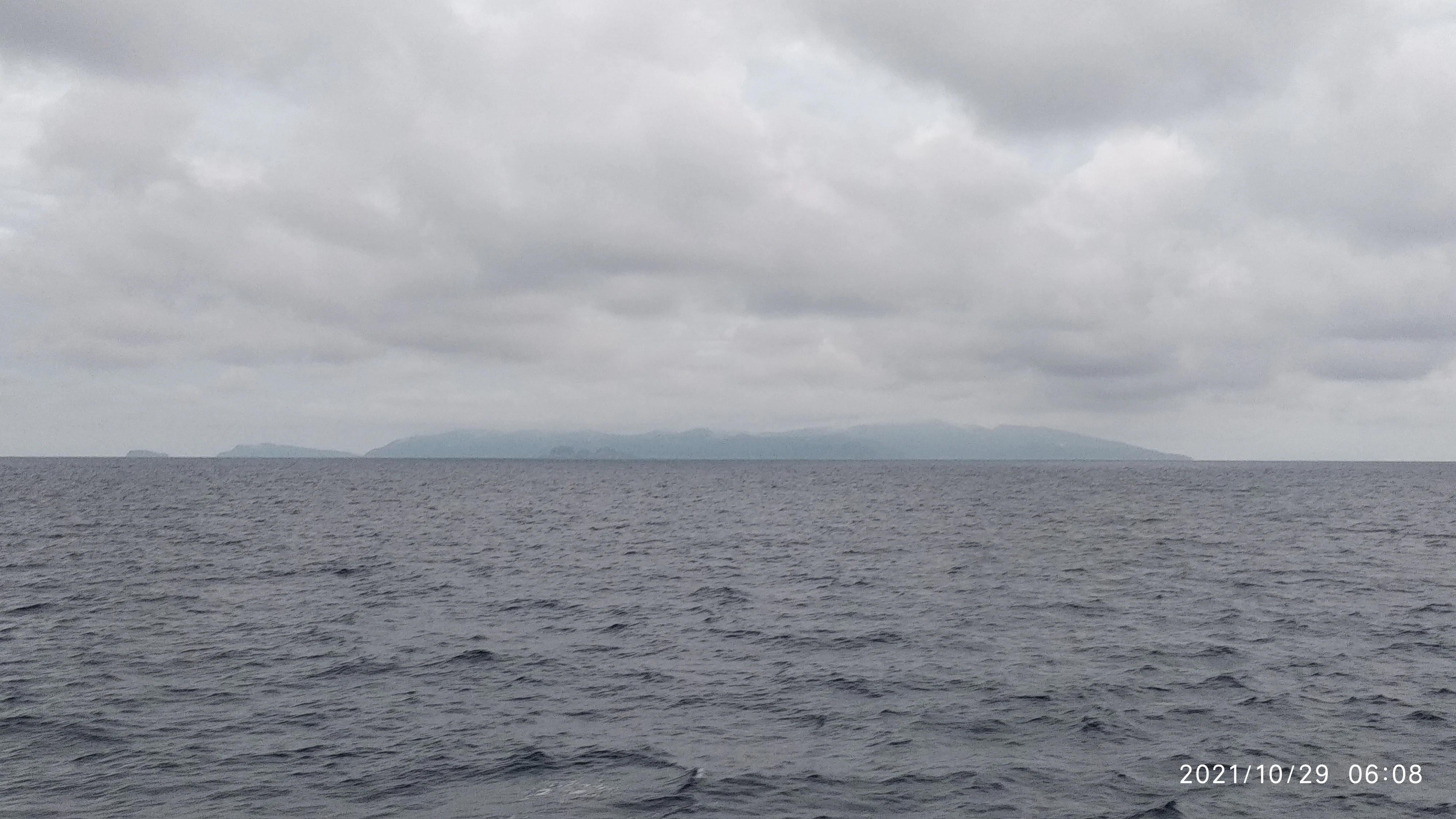 A distant, low-lying island silhouette on the horizon over calm, dark ocean water under a heavy, overcast sky.