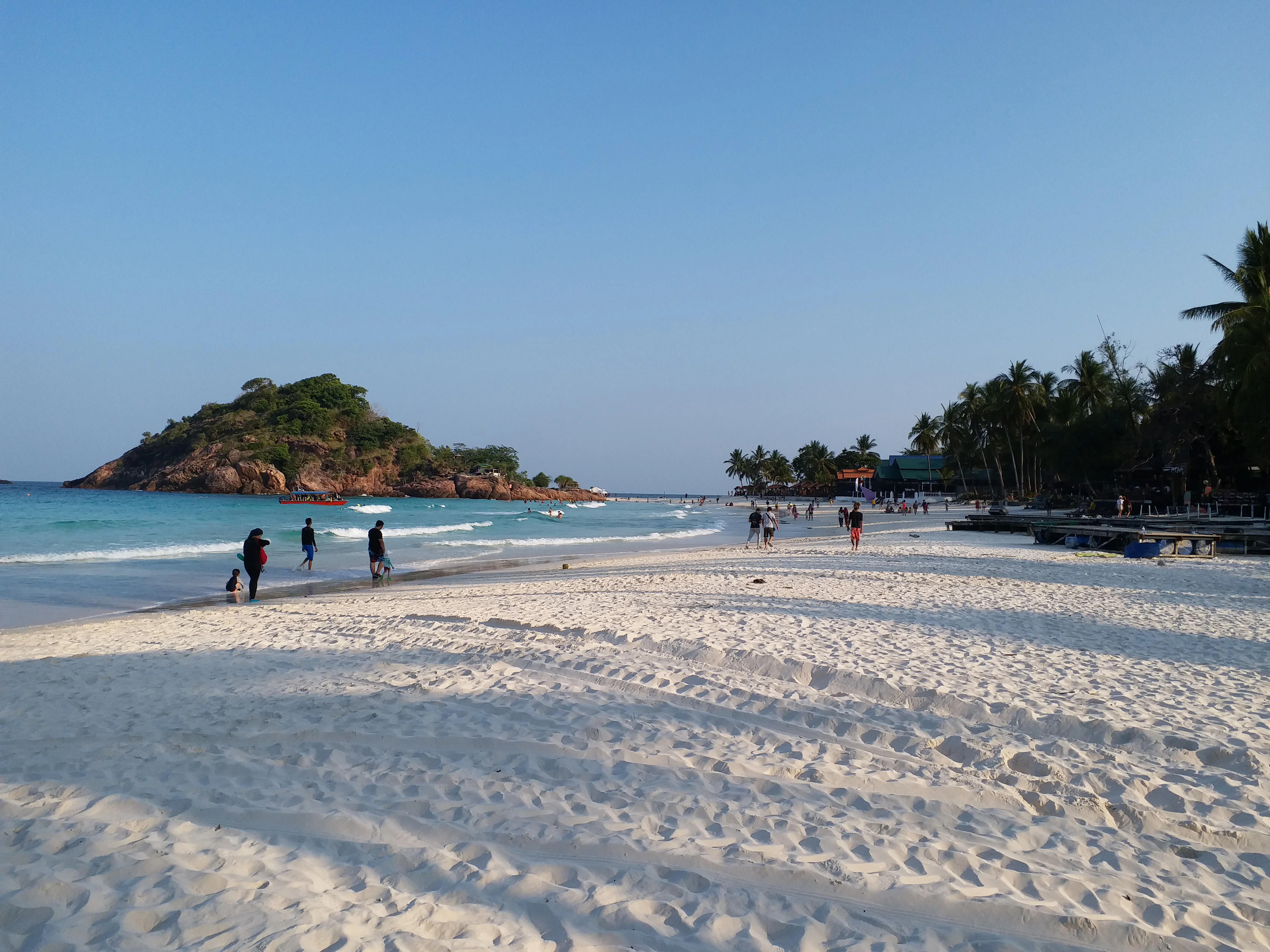 A wide, white sandy beach on Redang Island with turquoise water, a rocky islet in the distance, and people walking along the shore.