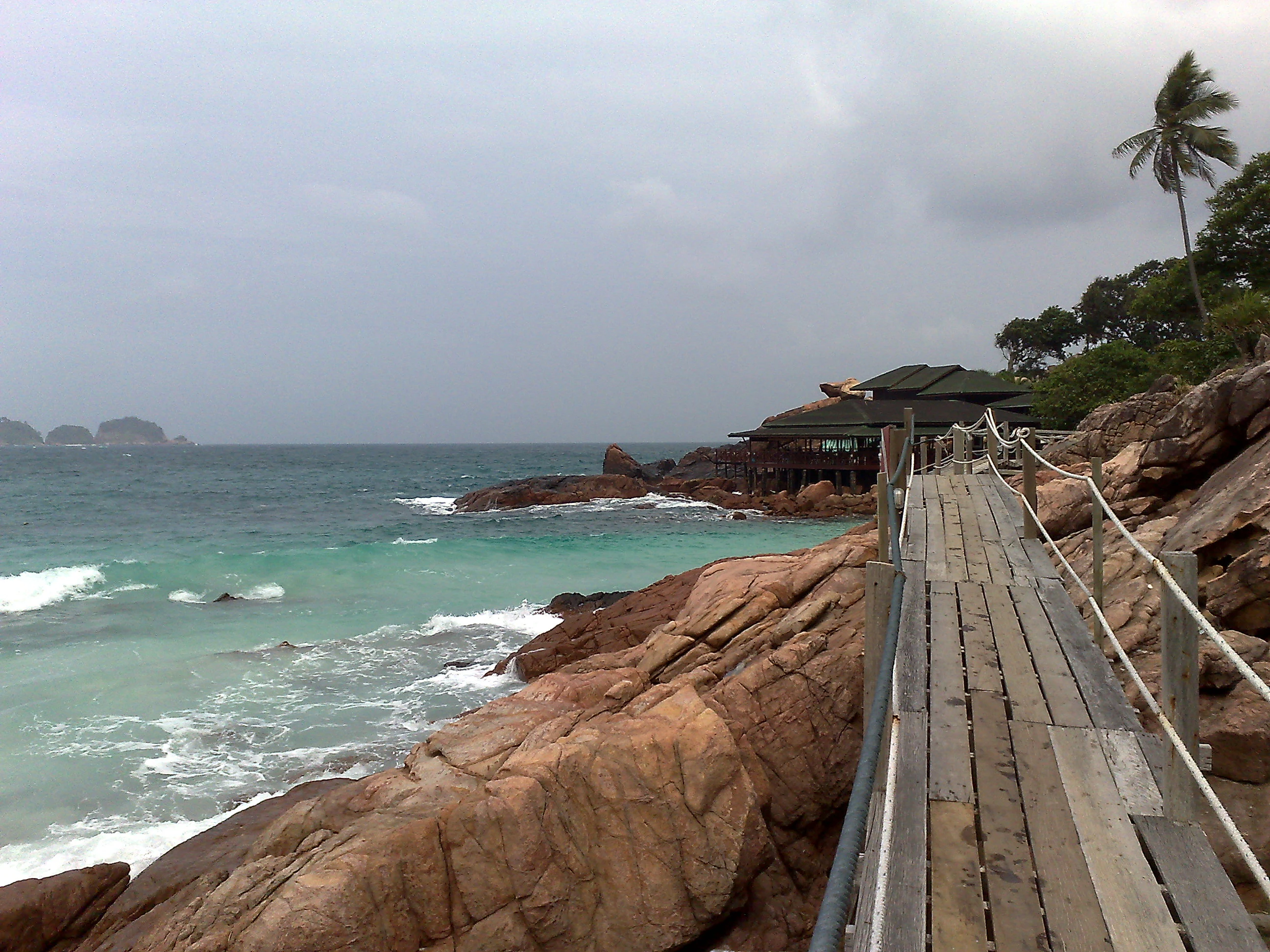 A weathered wooden footbridge path with a rope handrail, built into rocky terrain along a turquoise sea coast on Redang Island.