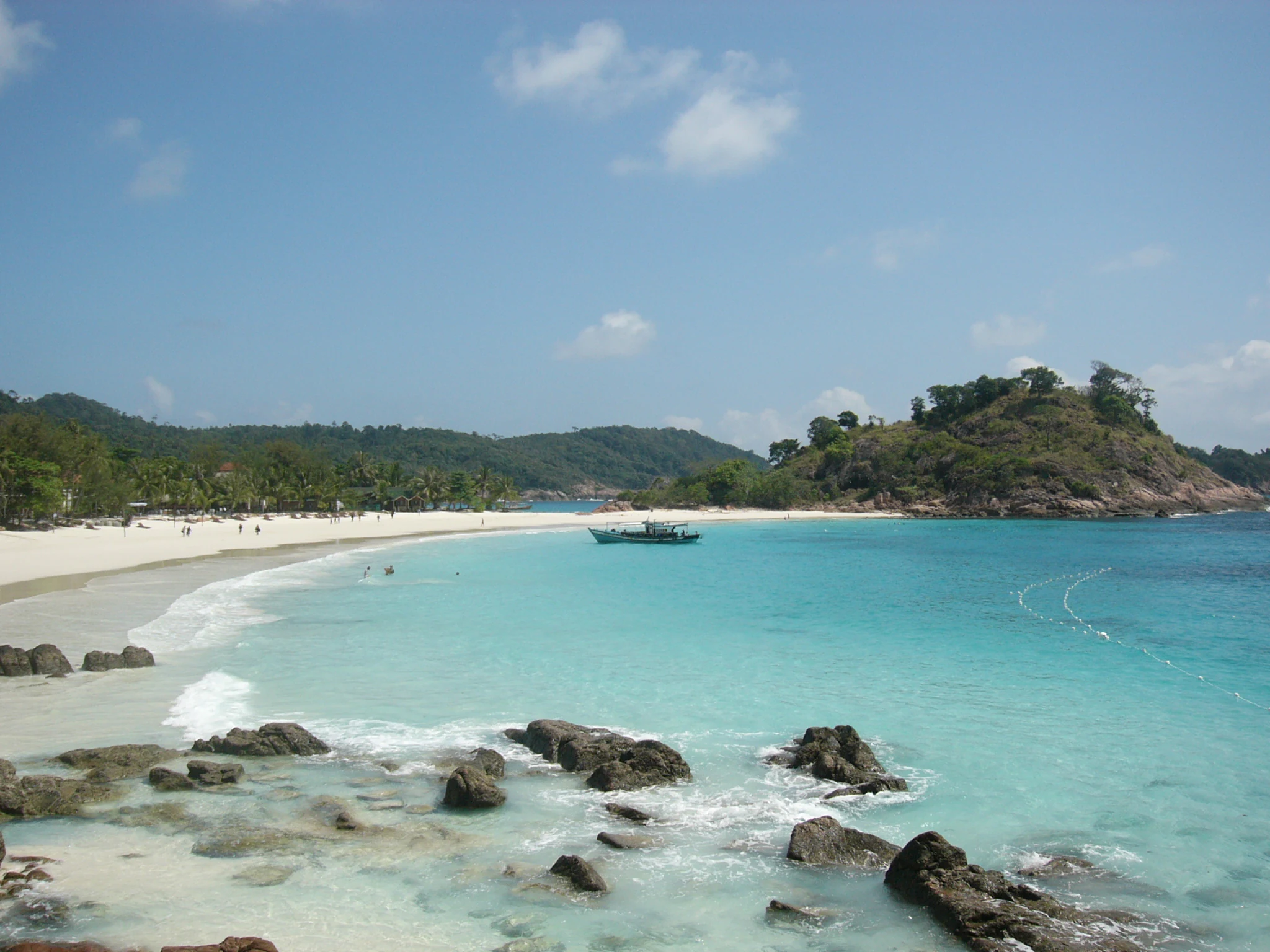 A wide angle view of a white sand beach meeting turquoise ocean waters on Redang Island with rocky formations in the foreground and hilly, forested headlands in the distance.