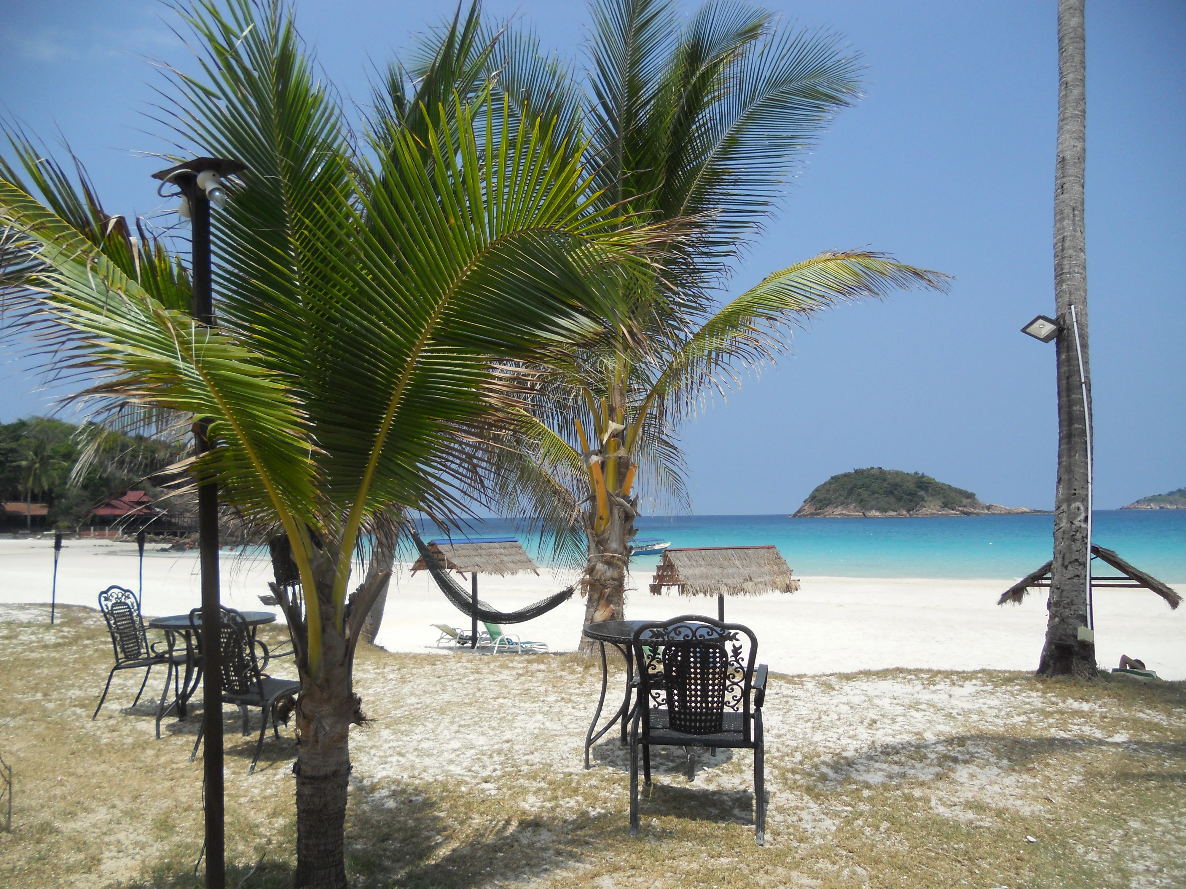 A sandy beach at Redang Island with palm trees, outdoor metal tables and chairs, and a hammock in the foreground, looking out toward a calm turquoise sea with small islands in the distance.