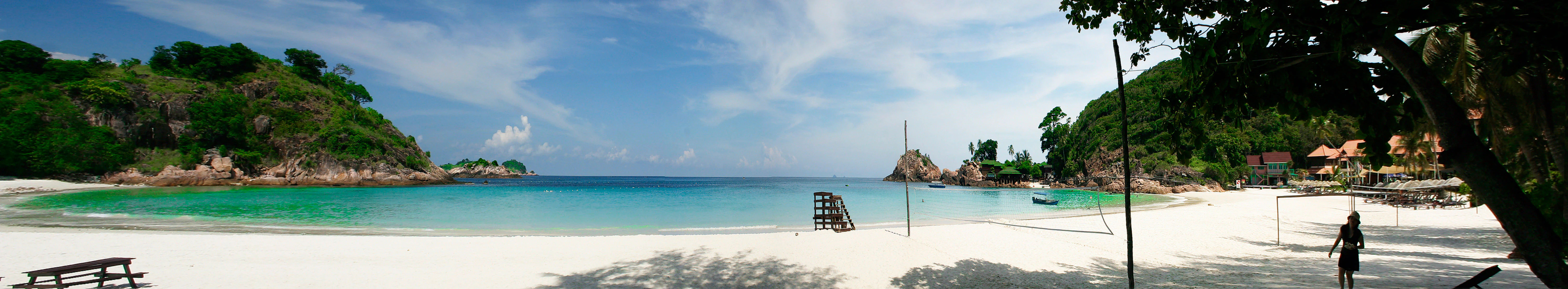 A wide panoramic view of a tropical beach with white sand, turquoise water, rocky green hills on both sides, and a few small structures visible near the shore.