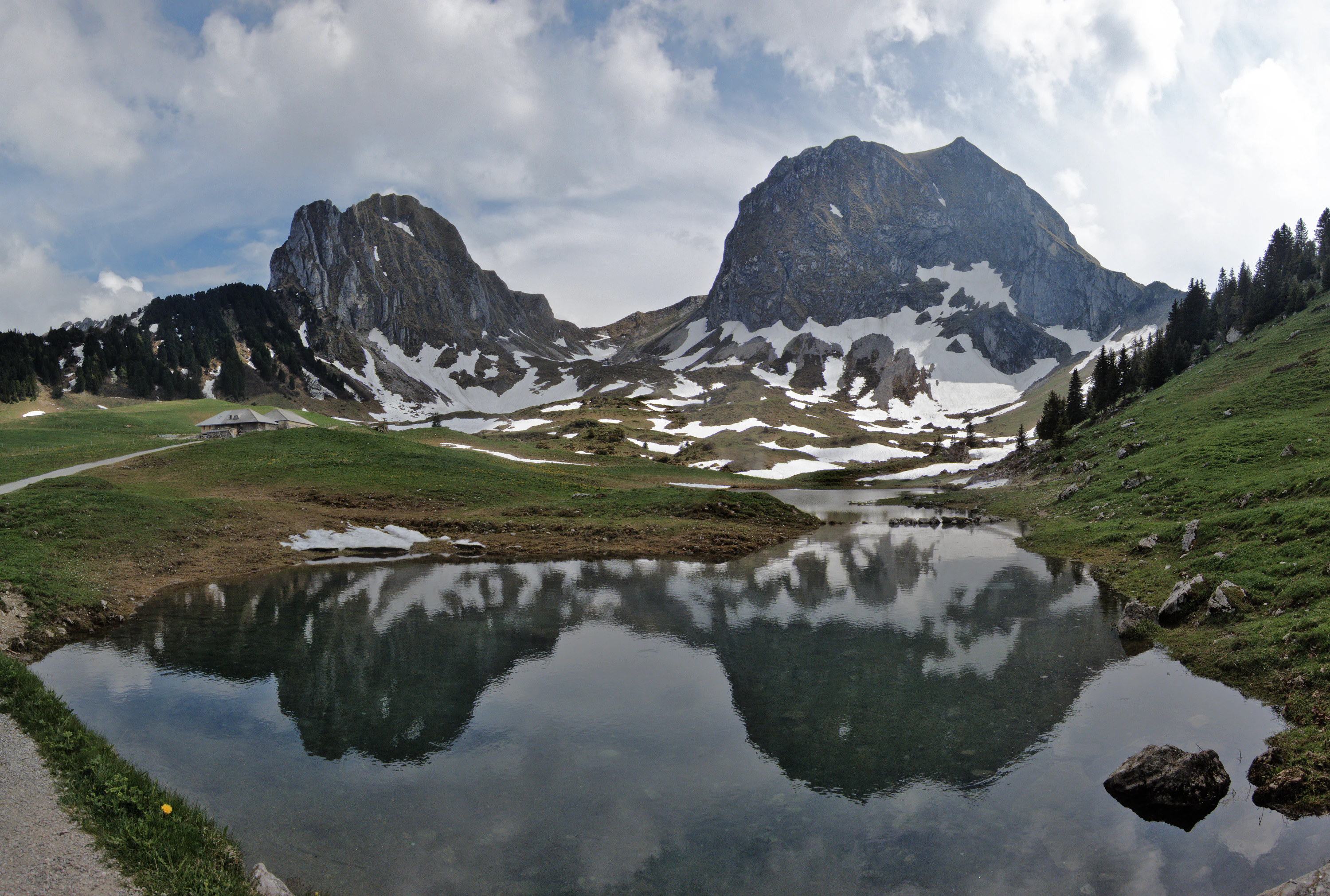 A wide angle view of the rugged Nüneneflue and Gantrisch mountain peaks partially covered in snow, reflected in a still alpine lake in the foreground.