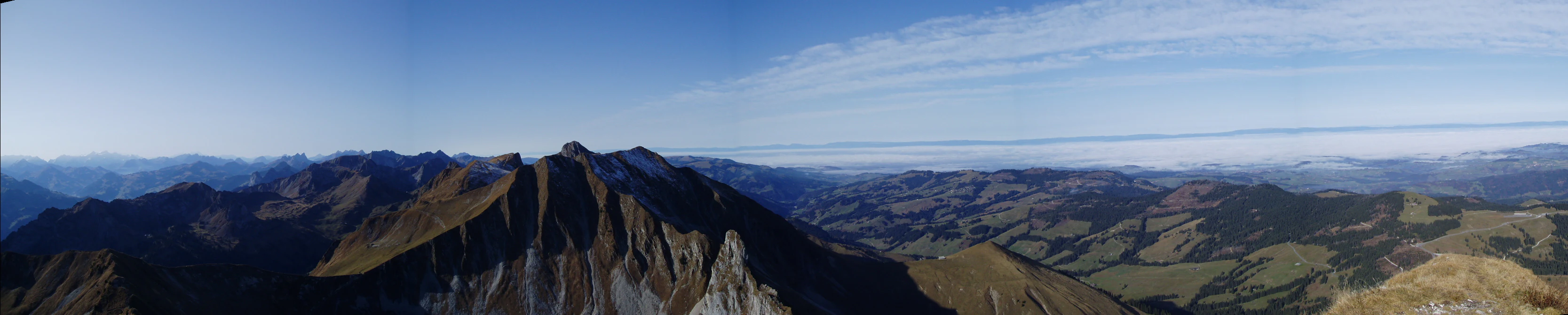 A wide panoramic mountain landscape under a clear blue sky, showing rugged ridgelines and rolling alpine terrain.