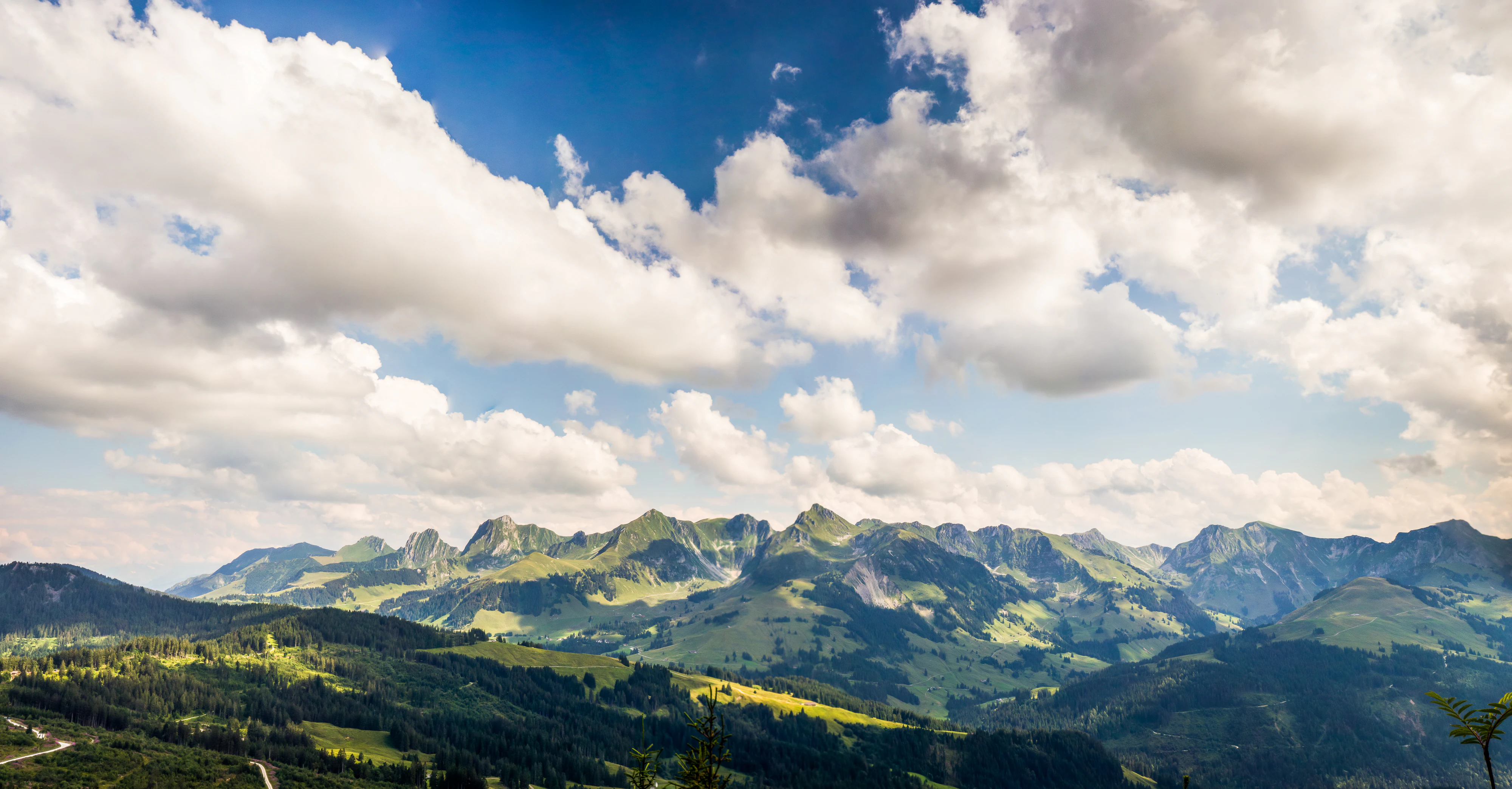 A wide, scenic view of a jagged, green mountain range under a blue sky with scattered white clouds.