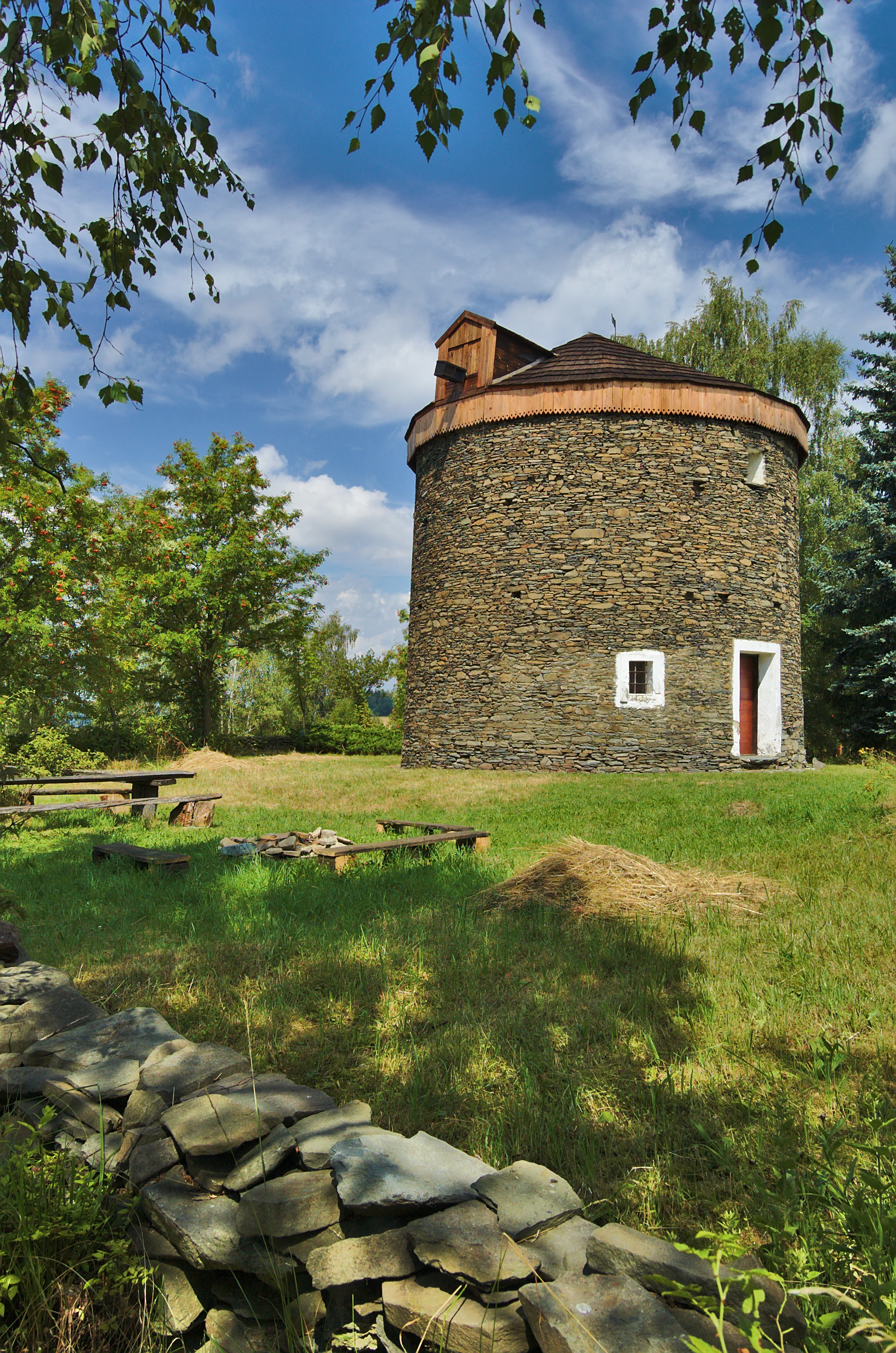 A historic circular stone windmill with a wooden roof stands in a grassy field on a sunny day, surrounded by trees and a stone wall in the foreground.