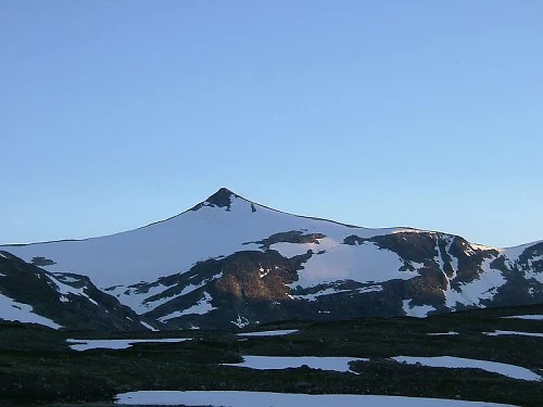 Mountain peak with snow patches on its slopes and rocky areas visible against a clear blue sky