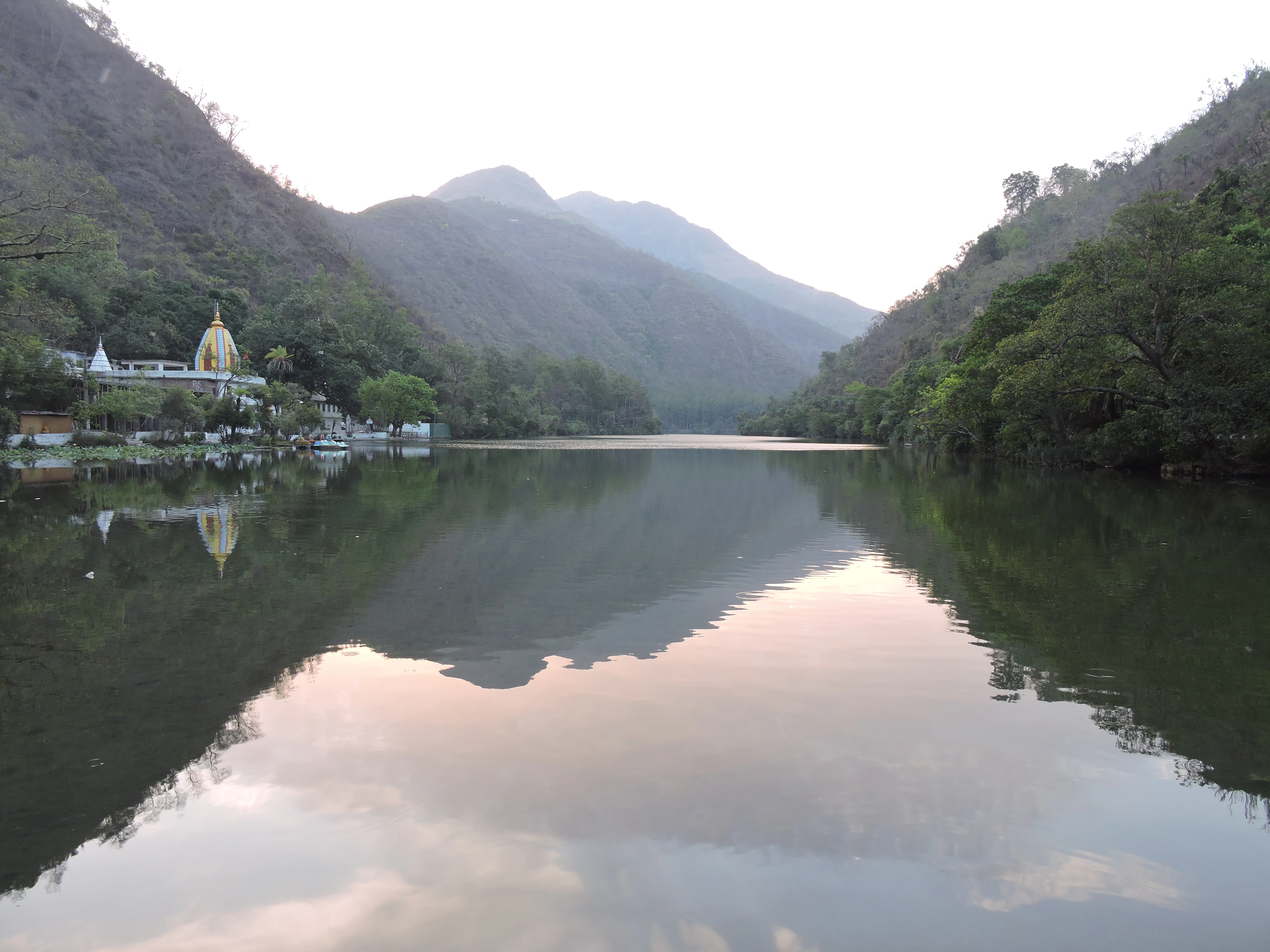 A wide view of a calm, reflective lake surrounded by forested mountains under a soft light, with a golden temple visible on the left shore.