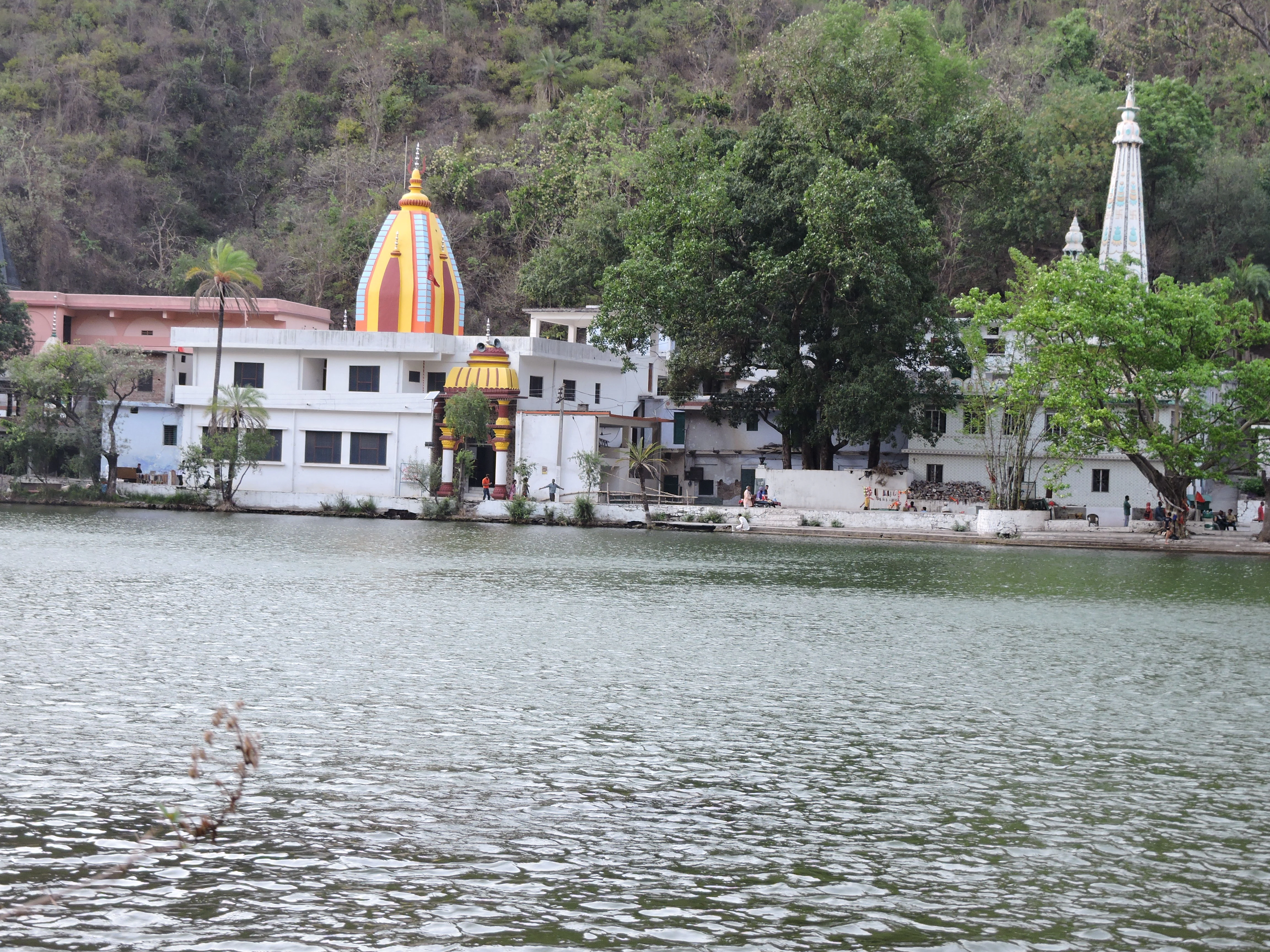 A white temple with a bright yellow and orange dome, and a tall, slender white spire, stands on the edge of a calm lake surrounded by trees and a forested hillside.