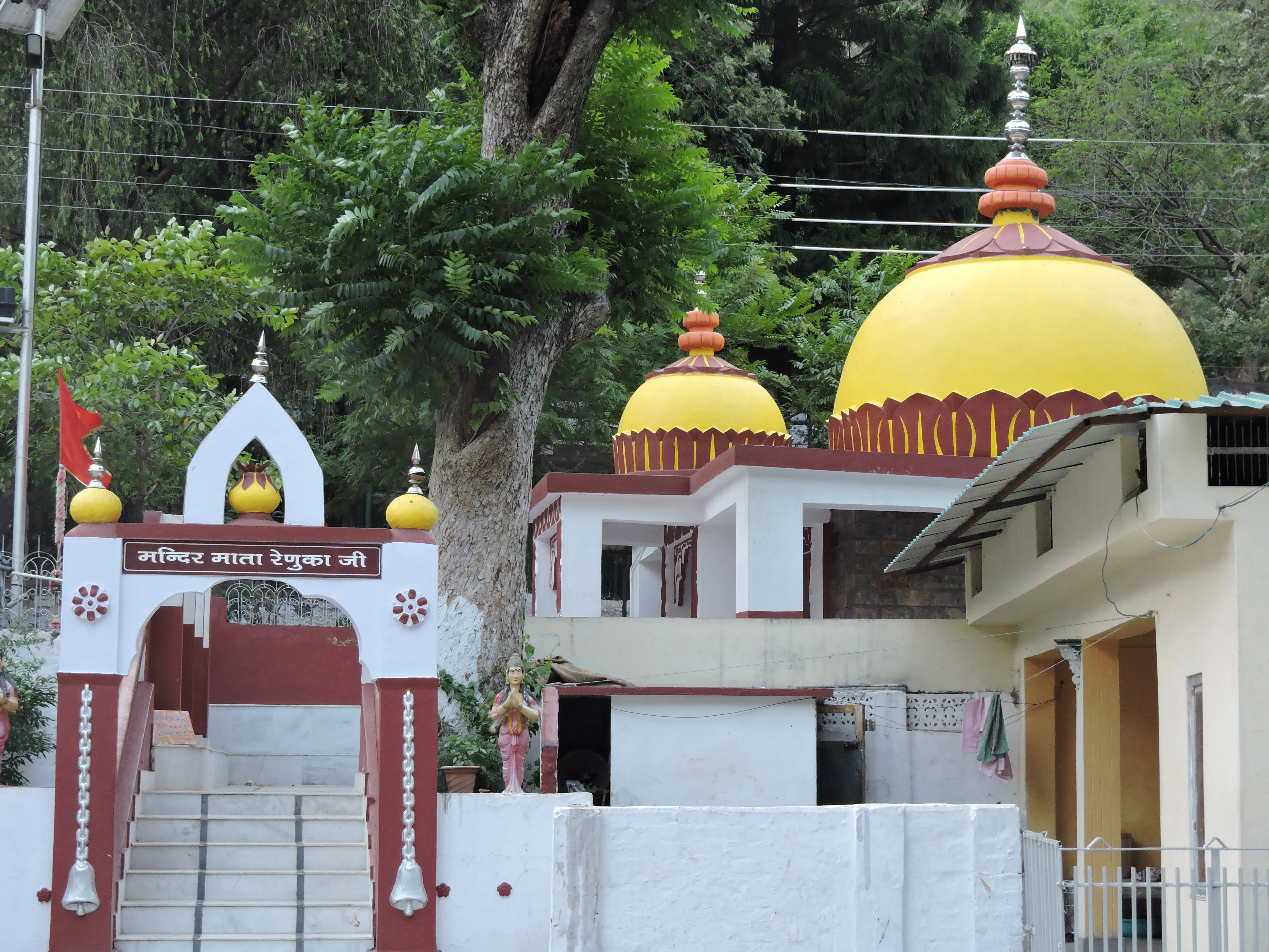 A view of the white and maroon entrance gate to the Mata Renuka Ji Temple with bright yellow domes in the background against a backdrop of green trees.
