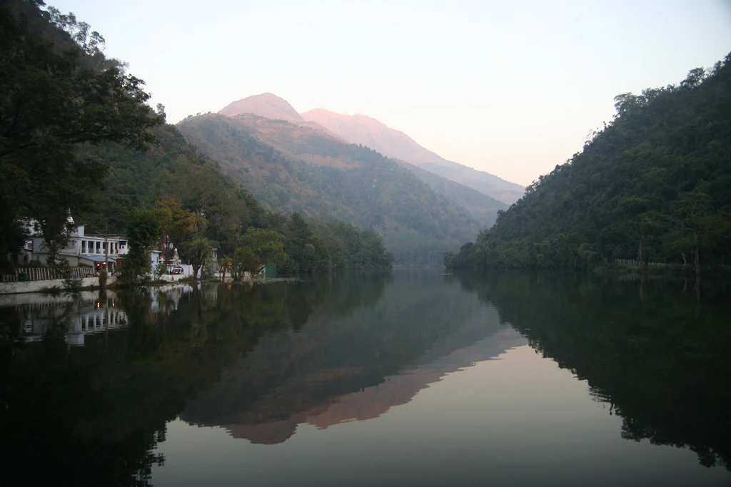 A calm lake reflects the image of surrounding green mountain slopes and a small white structure on the shore.