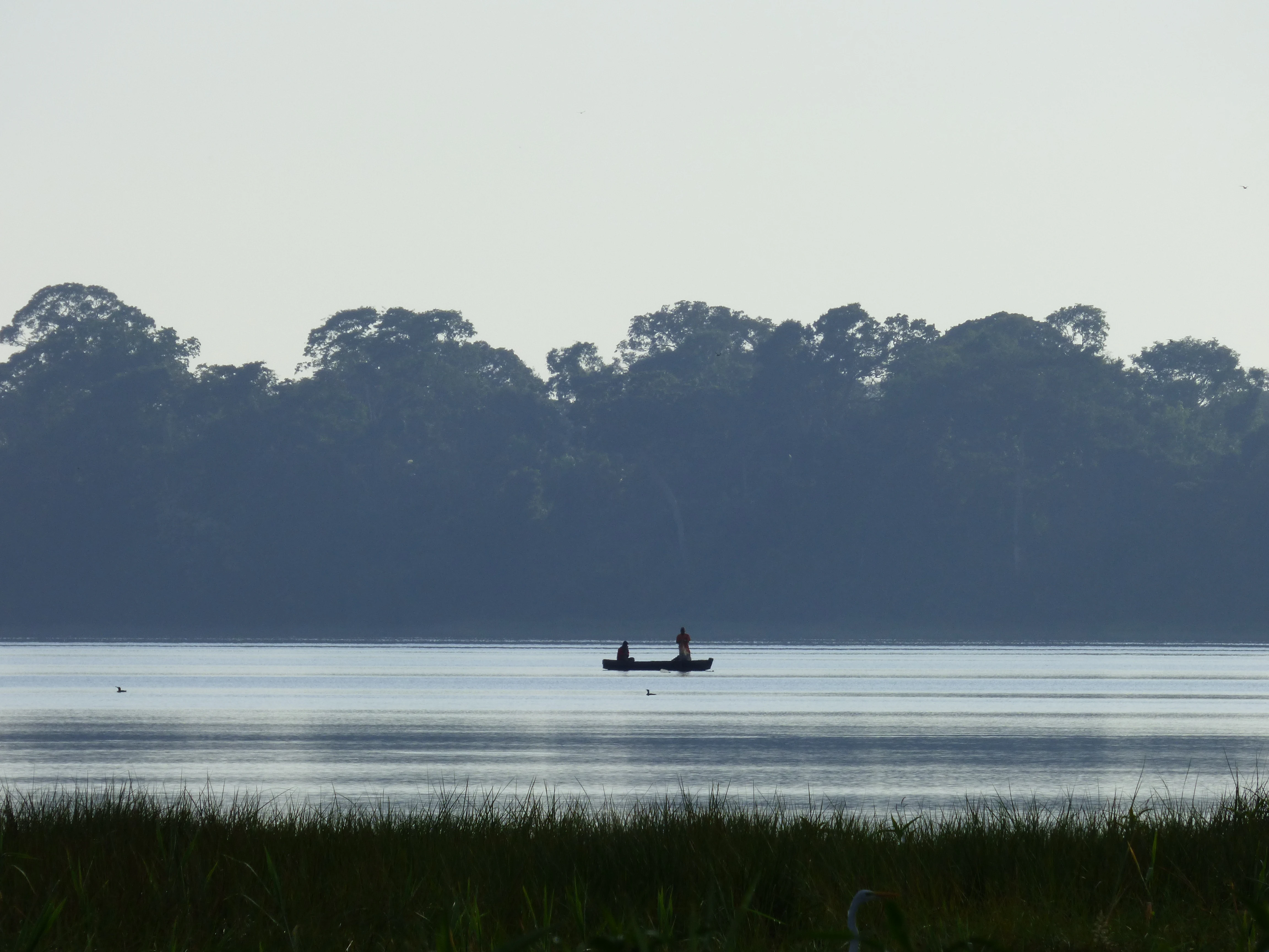 A small boat with two people fishing on a calm lake, silhouetted against a backdrop of dense forest and a light, misty sky at sunrise.