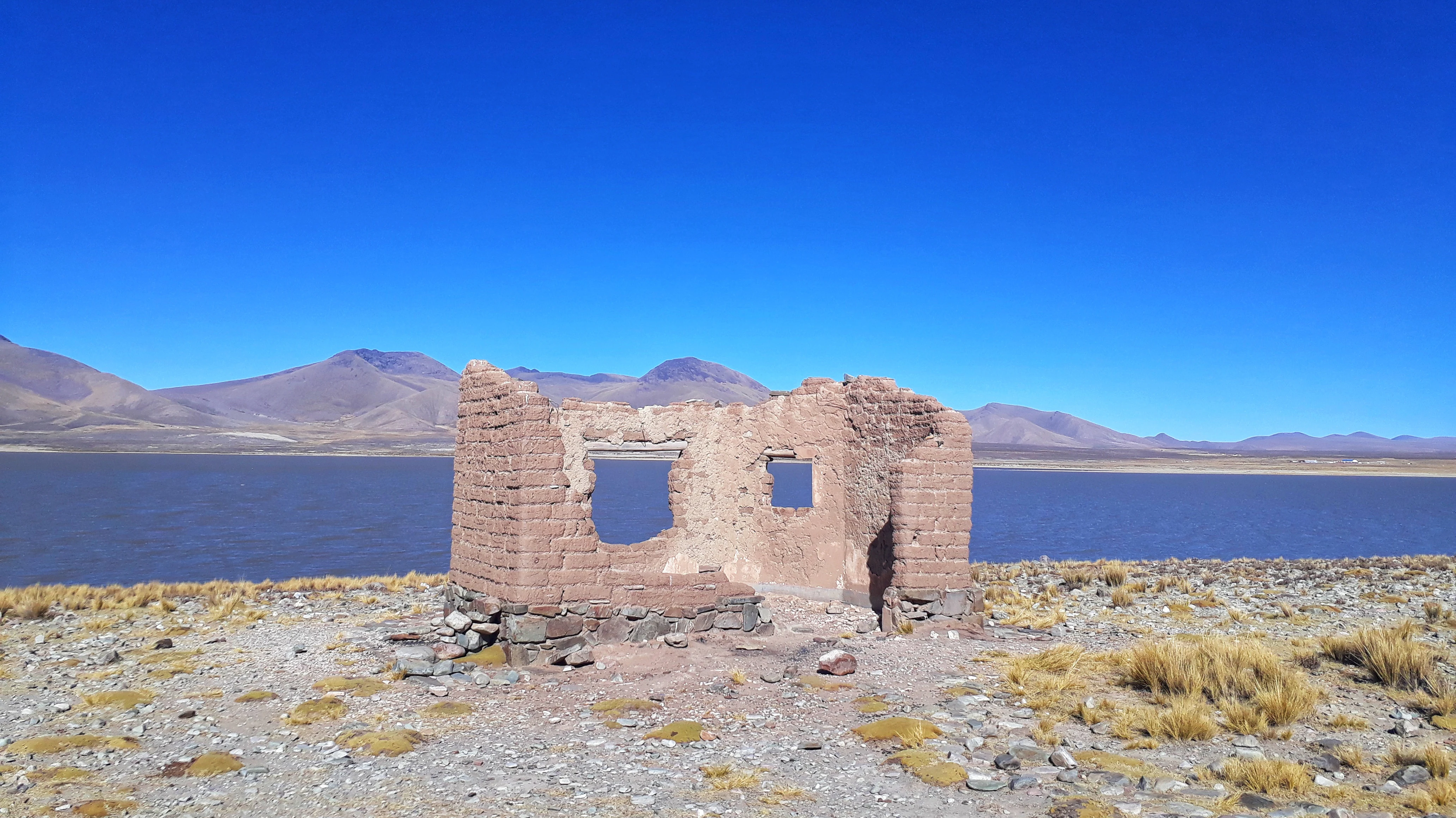 Una estructura de adobe en ruinas con dos aberturas de ventana y una entrada se alza sobre una orilla rocosa y cubierta de hierba frente a una gran laguna azul, con montañas a lo lejos bajo un cielo azul despejado.