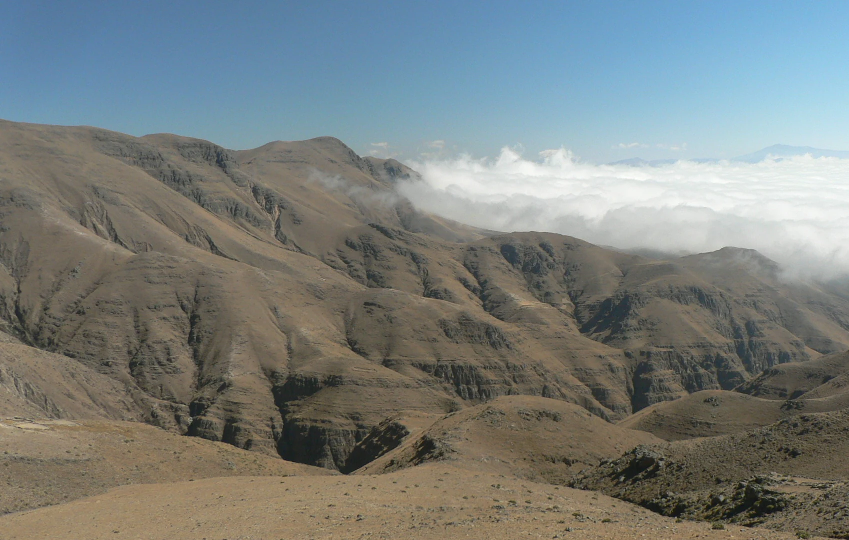 Vista panorámica de crestas montañosas secas y pardas, con valles profundos y una capa de nubes blancas bajo las cumbres, todo ello bajo un cielo azul despejado.