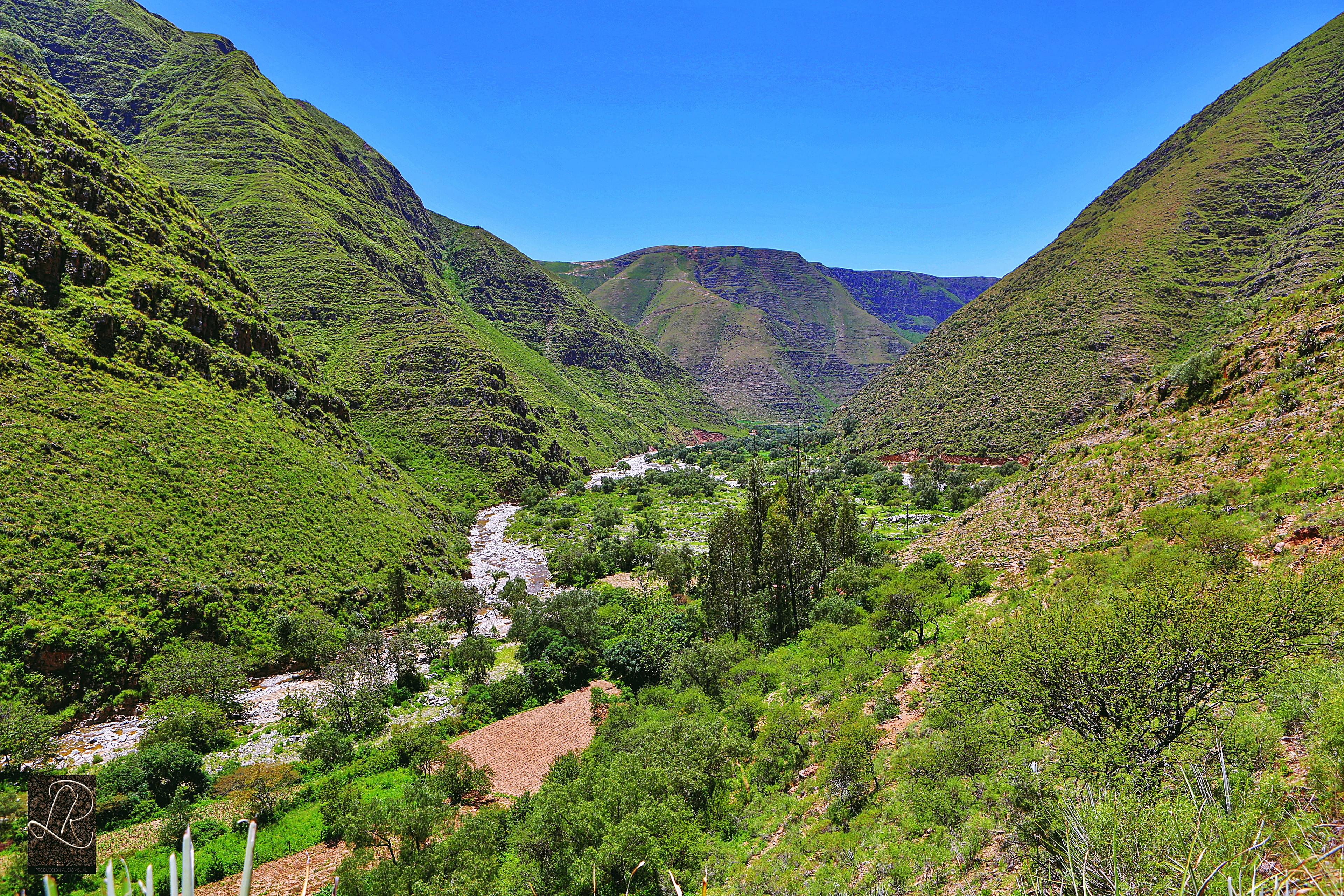 Plano general de un valle de montaña verde con un río que fluye a través de él, rodeado de laderas escarpadas cubiertas de vegetación, con un pequeño pueblo visible en la distancia bajo un cielo azul despejado.