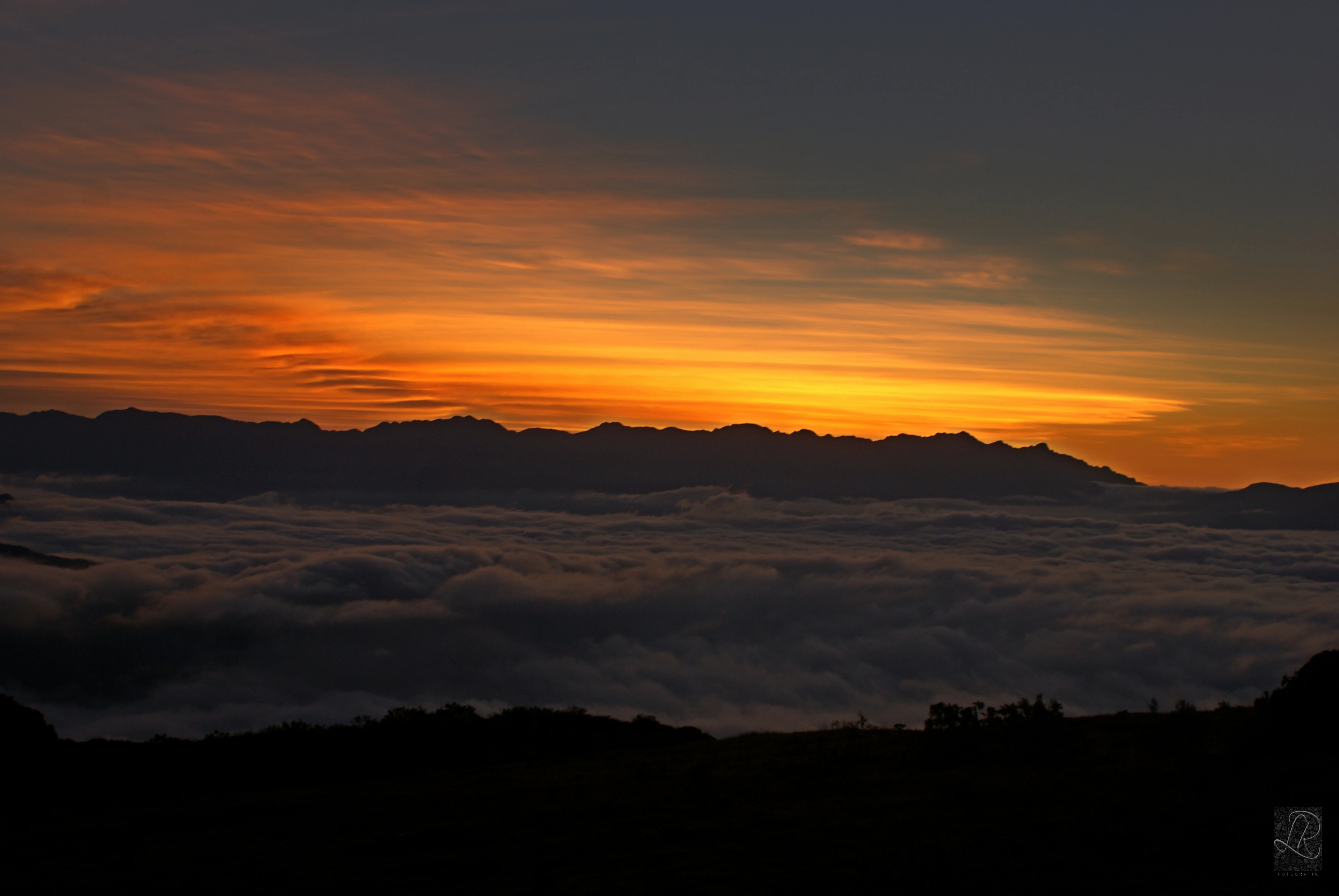 Una fotografía de paisaje que muestra un cielo al amanecer con tonos vibrantes naranja y amarillo sobre una cordillera silueteada, con una densa capa de nubes o niebla en el valle inferior y un primer plano oscuro.