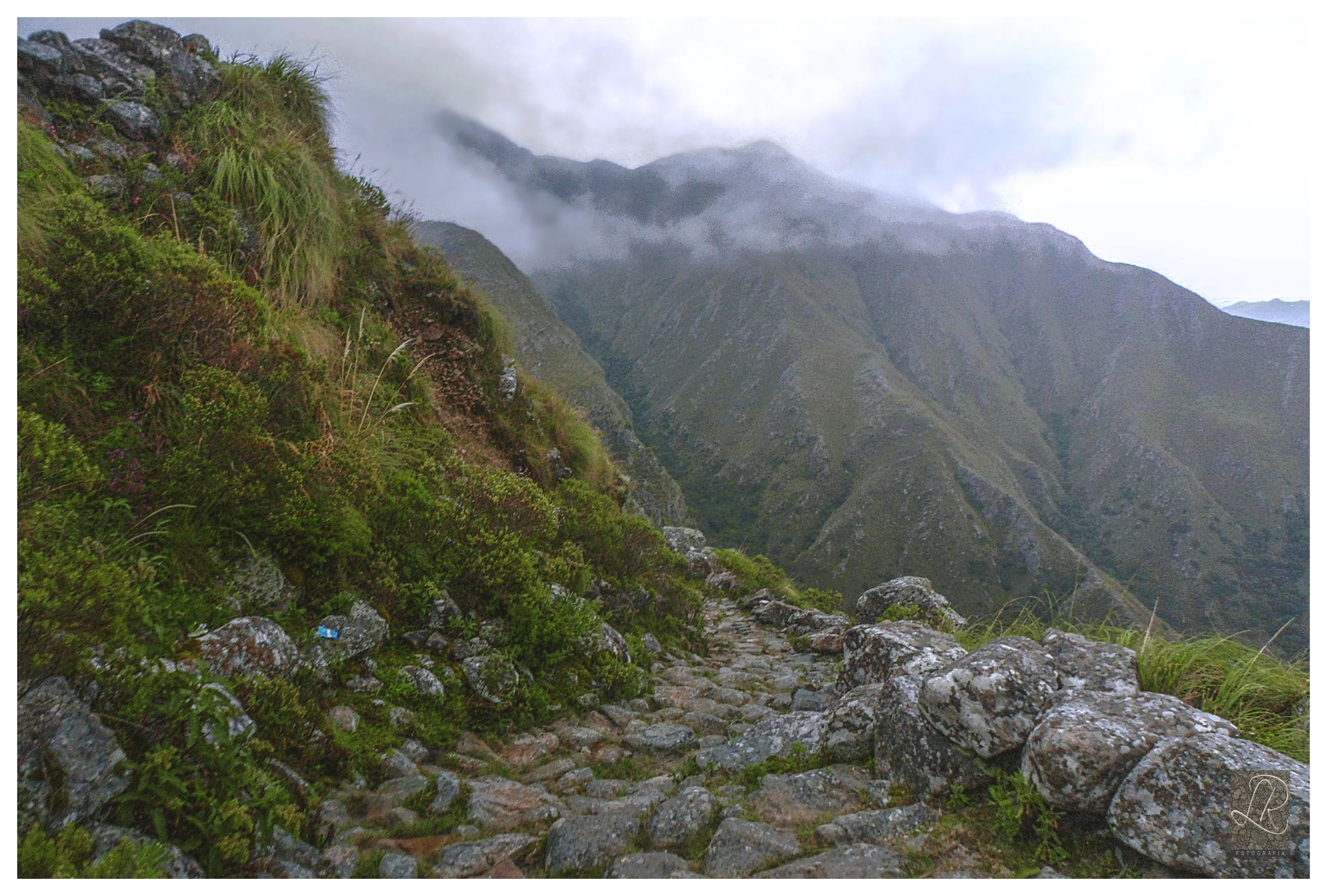 Un sendero pavimentado con piedra serpentea a lo largo del borde de una ladera de montaña empinada y herbosa, con montañas verdes y brumosas visibles al fondo.