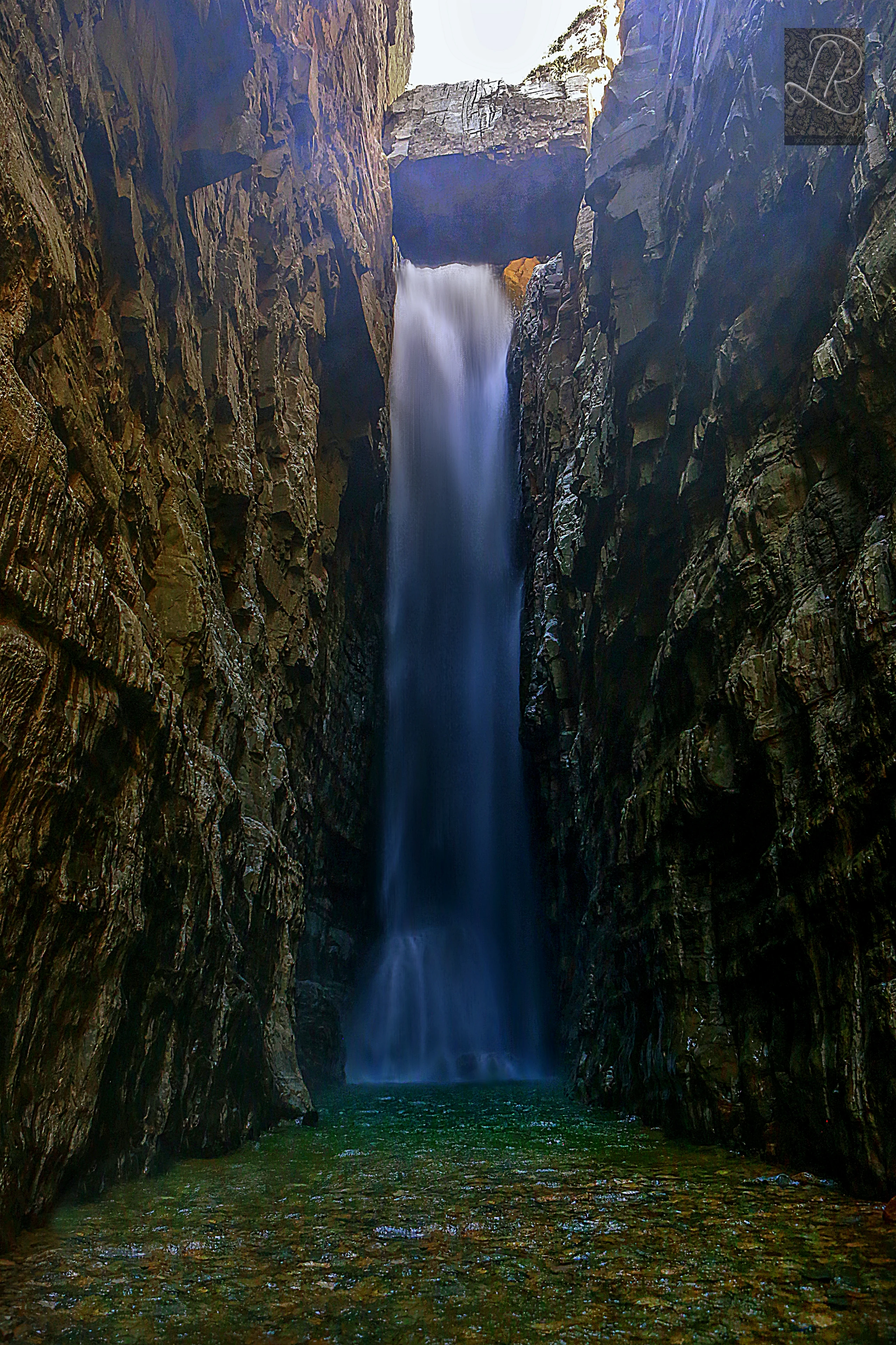 Una cascada alta que fluye a través de un estrecho cañón de roca con paredes escarpadas hacia una poza de tonos verdes, capturada con larga exposición para dar al agua un aspecto sedoso y suave