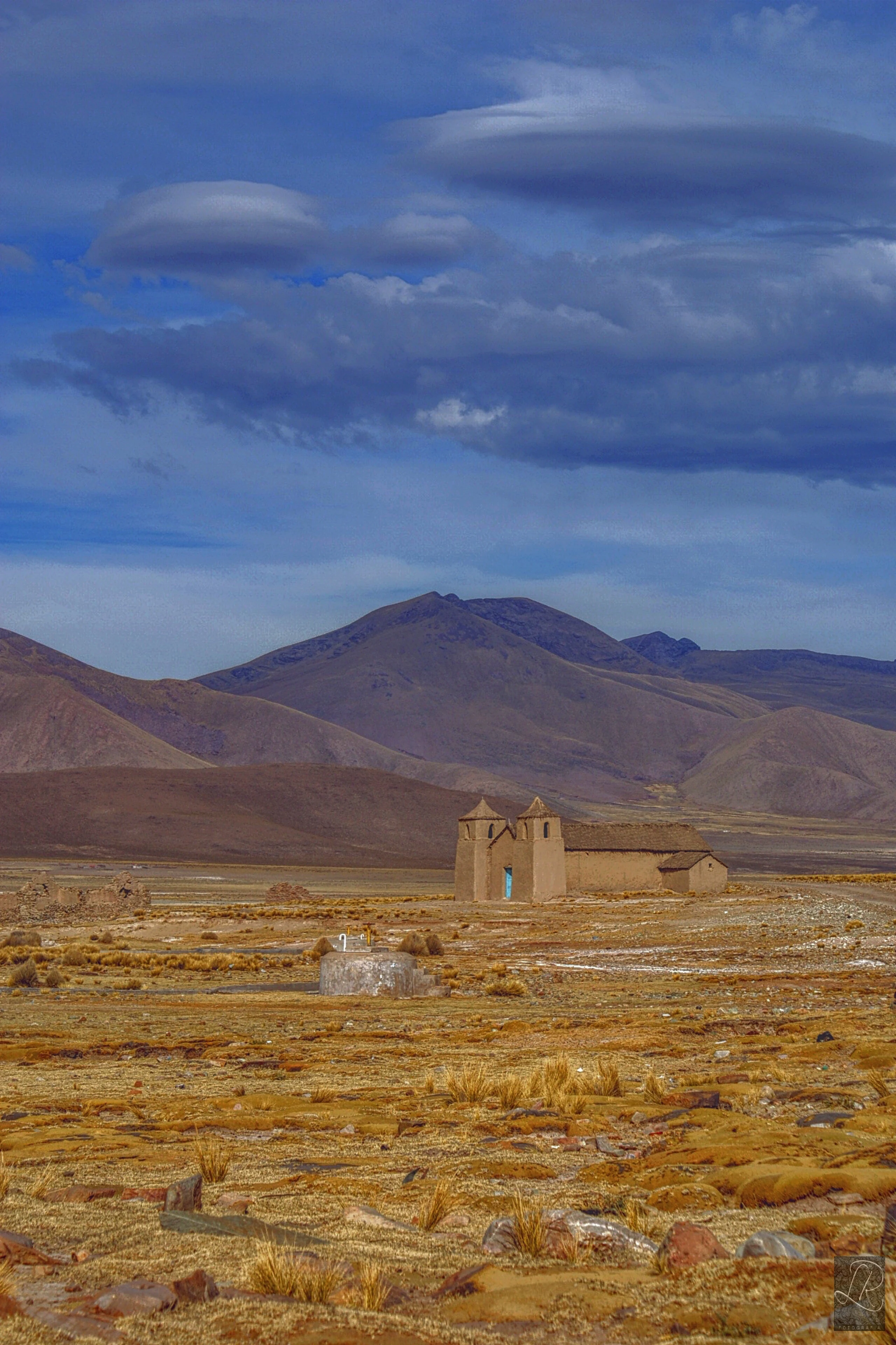 Una pequeña iglesia de adobe con dos torres campanario se alza en una vasta llanura seca y rocosa con escasa hierba amarillenta, flanqueada por montañas marrones bajo un cielo azul espectacular con nubes oscuras.