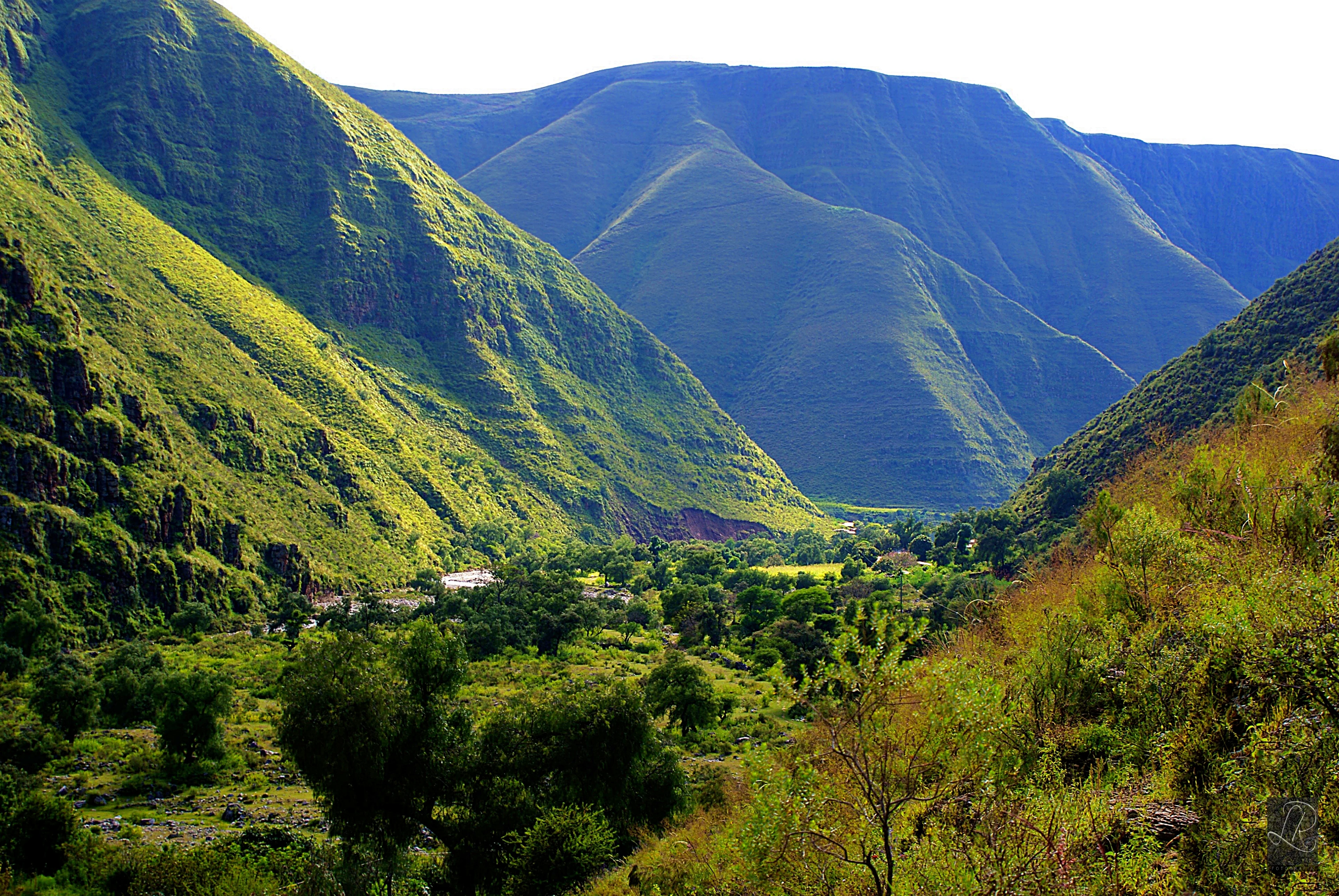 Fotografía panorámica de un valle montañoso verde con laderas empinadas y un pequeño grupo de edificios a lo lejos.