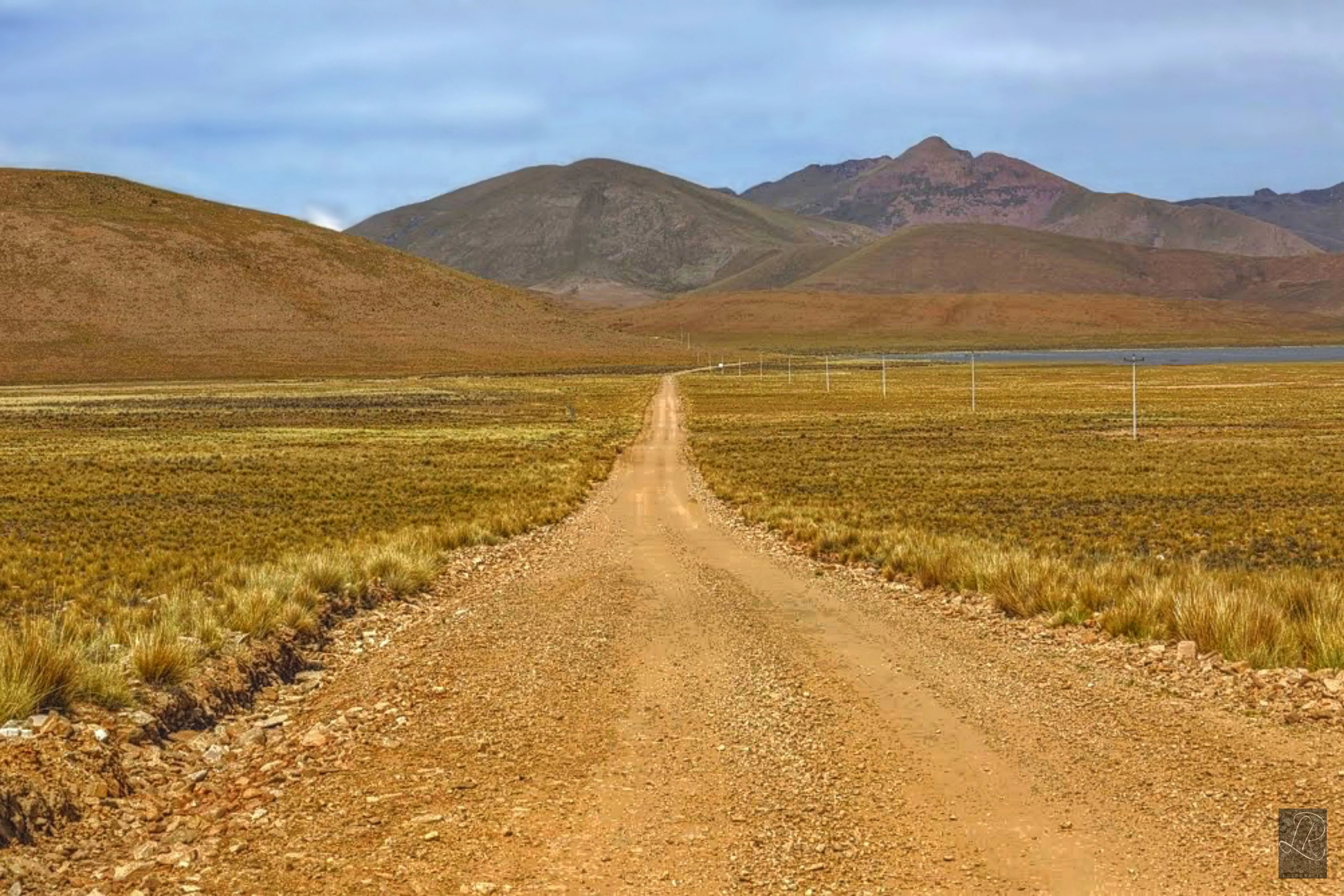 Un camino de grava recto se extiende a través de una pradera abierta hacia montañas distantes, con un cuerpo de agua visible a la derecha y una cerca a lo largo del camino