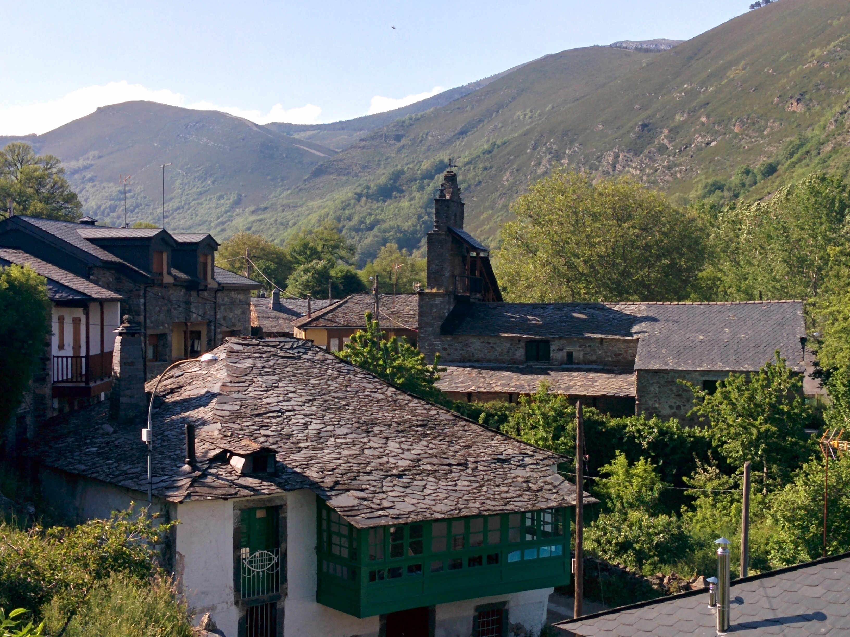 A cluster of stone houses with dark slate roofs sits in a lush green valley, backed by steep, grassy mountainsides under a clear sky.