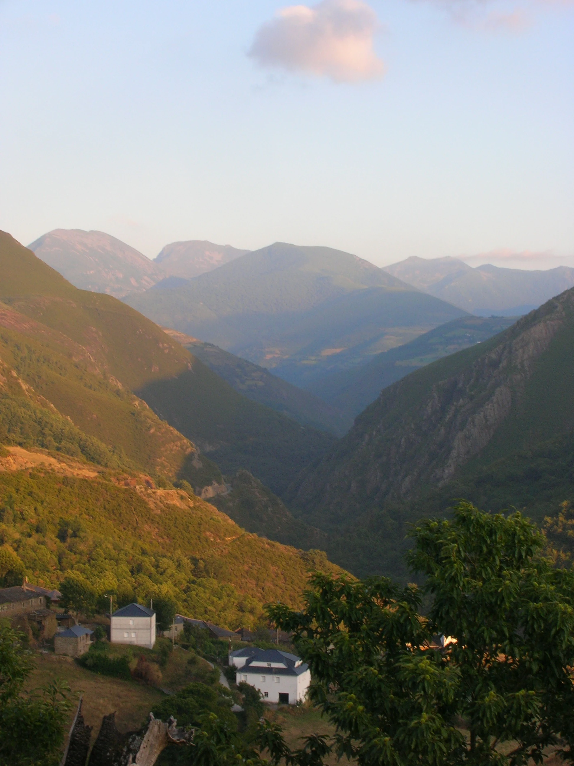 Una vista gran angular de un valle montañoso verde y frondoso con pequeñas casas de piedra en primer plano y capas de picos montañosos que se desvanecen en la distancia bajo un cielo pálido.